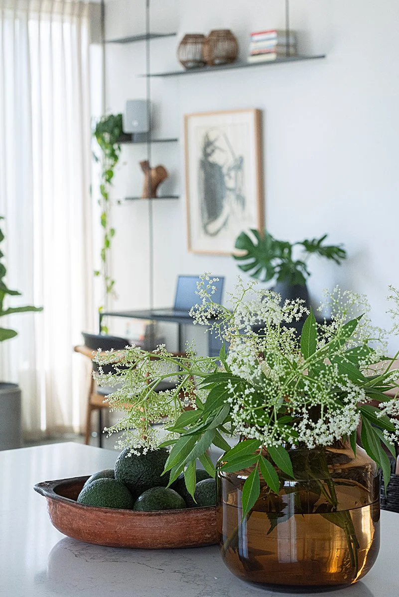 A glass vase with white flowers and green leaves on a white table, with a bowl of avocados nearby.