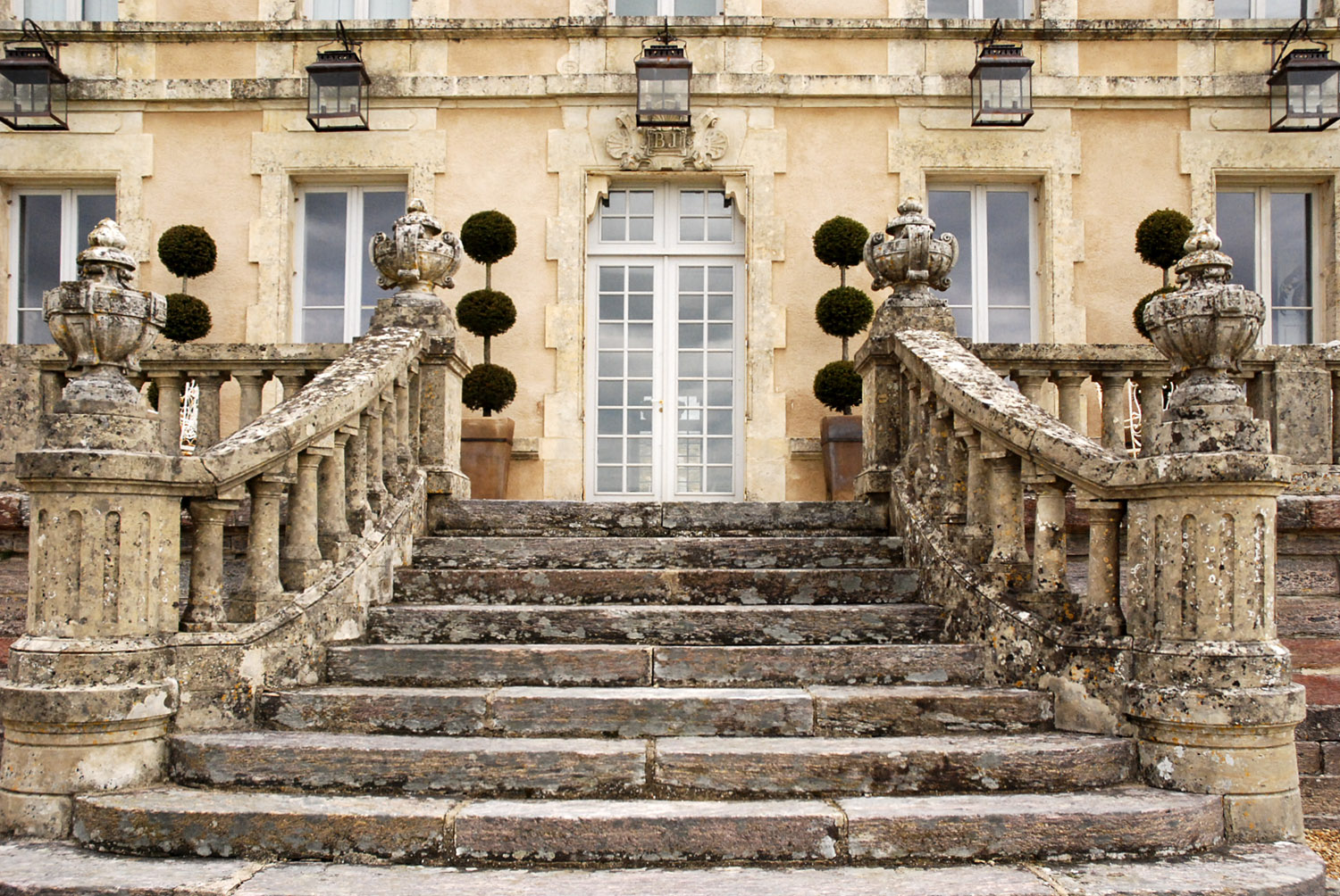 Stone staircase leading to a large ornate door with windowpanes. The staircase has decorative balustrades and urn-shaped finials, with potted topiary trees on either side.