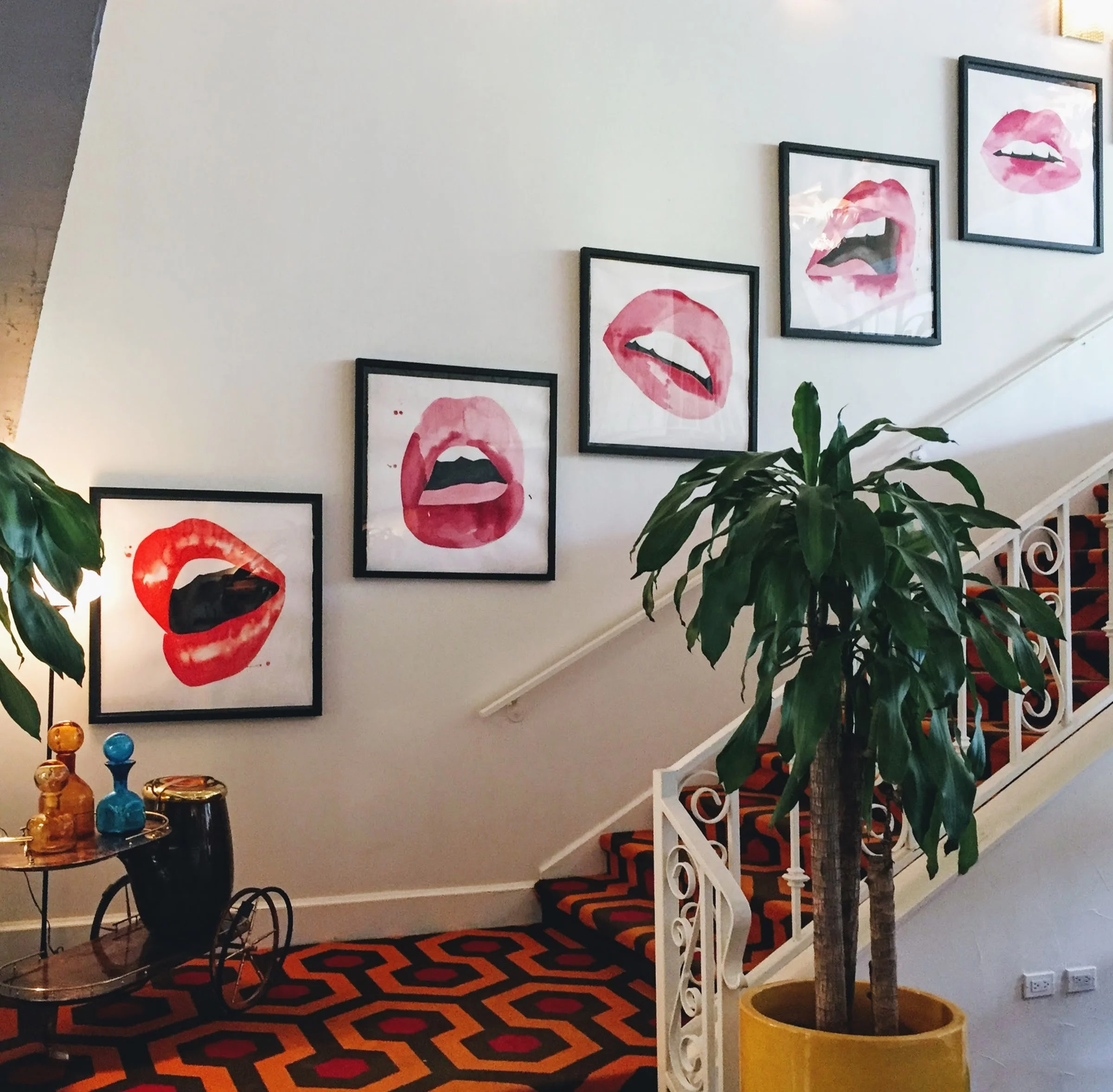Interior of a house staircase decorated with five framed watercolor art prints of open mouth lips, arranged diagonally along the wall. A large potted plant with lush green leaves stands next to the staircase, which has a colorful geometric patterned