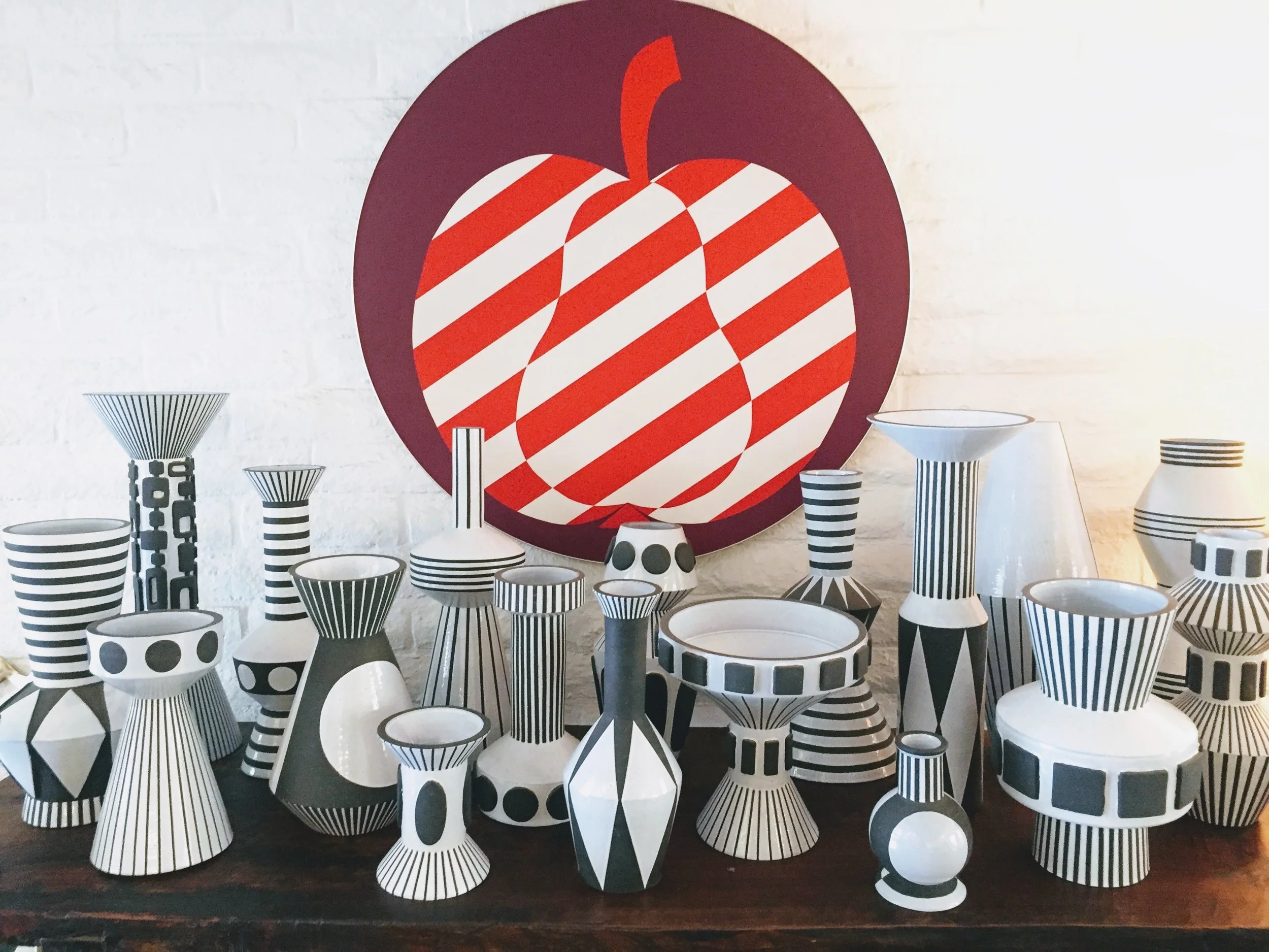 Collection of black and white patterned vases and bowls displayed on a wooden surface in front of a white brick wall with a large artwork of a strawberry in red, white, and purple colors.