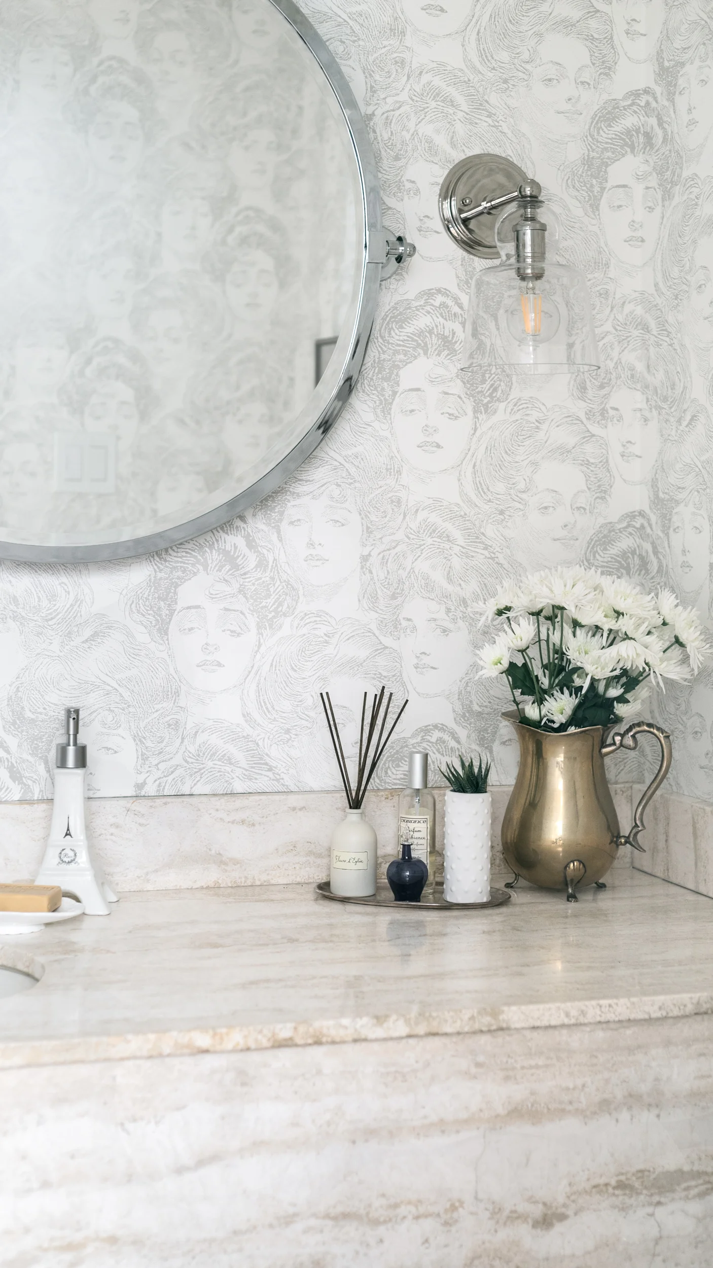 Bathroom counter with a vase of white flowers, small decorative bottles, a reed diffuser, and a soap dispenser. Reflection in the round mirror shows a patterned wall with illustrated women's faces.