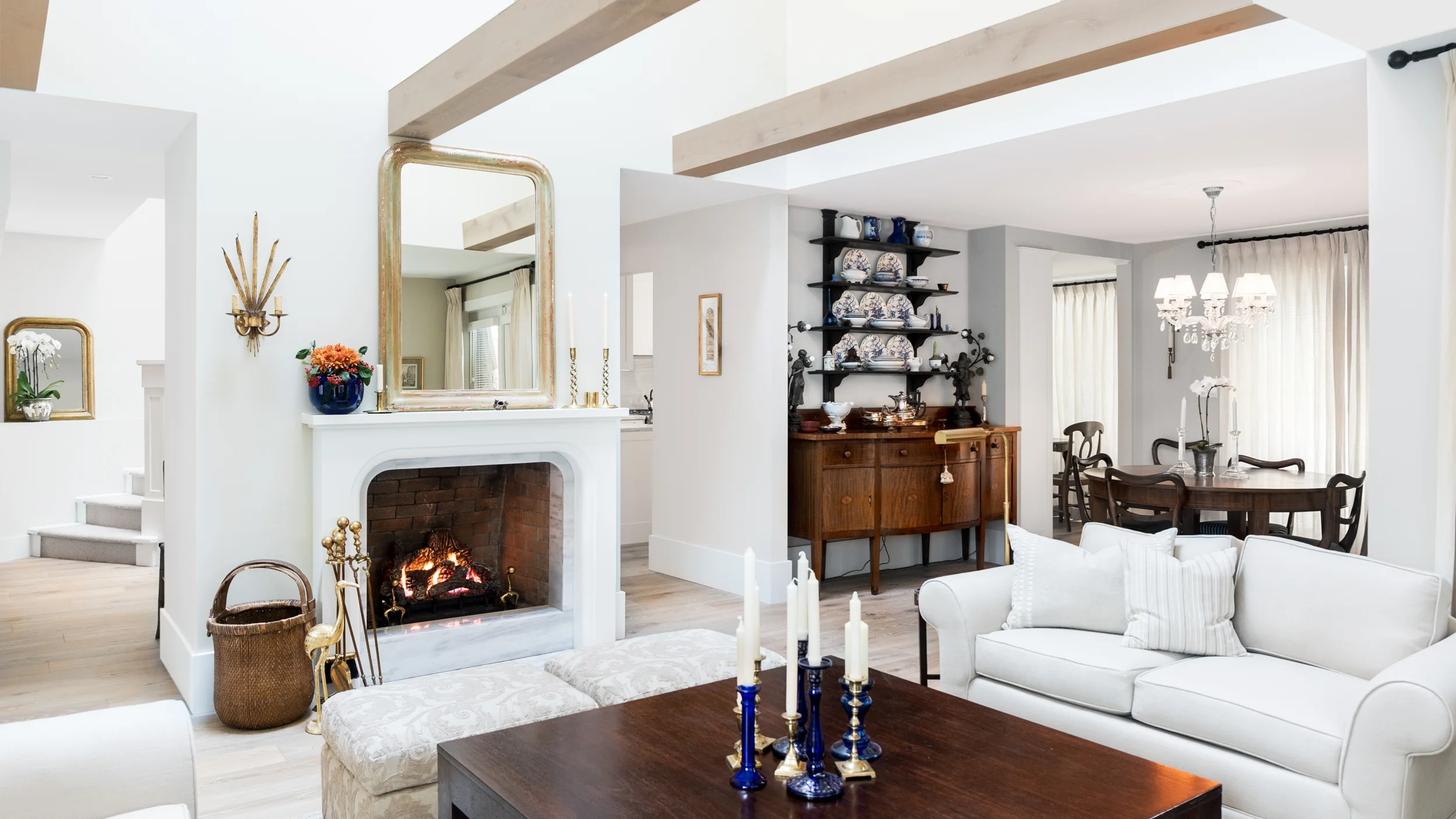 Living room with a white sofa, a fireplace with a mirror above it, a table with blue and white candlesticks, a wooden dining table with chairs, and a chandelier. Open floor plan with visible staircase and decorative shelves.