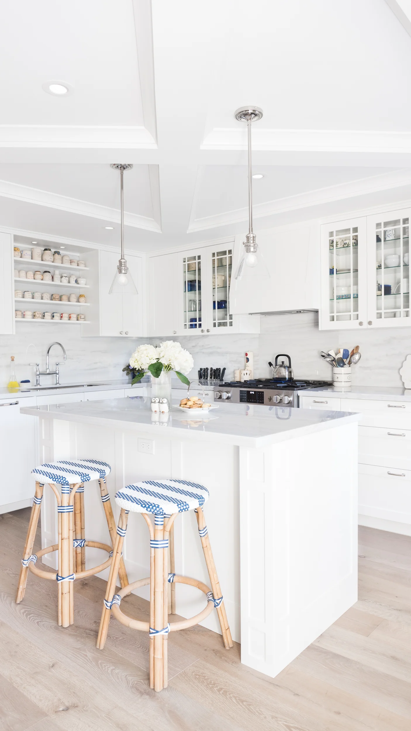 Bright white kitchen with island, two bar stools with blue and white cushions, flowers, and kitchen appliances.