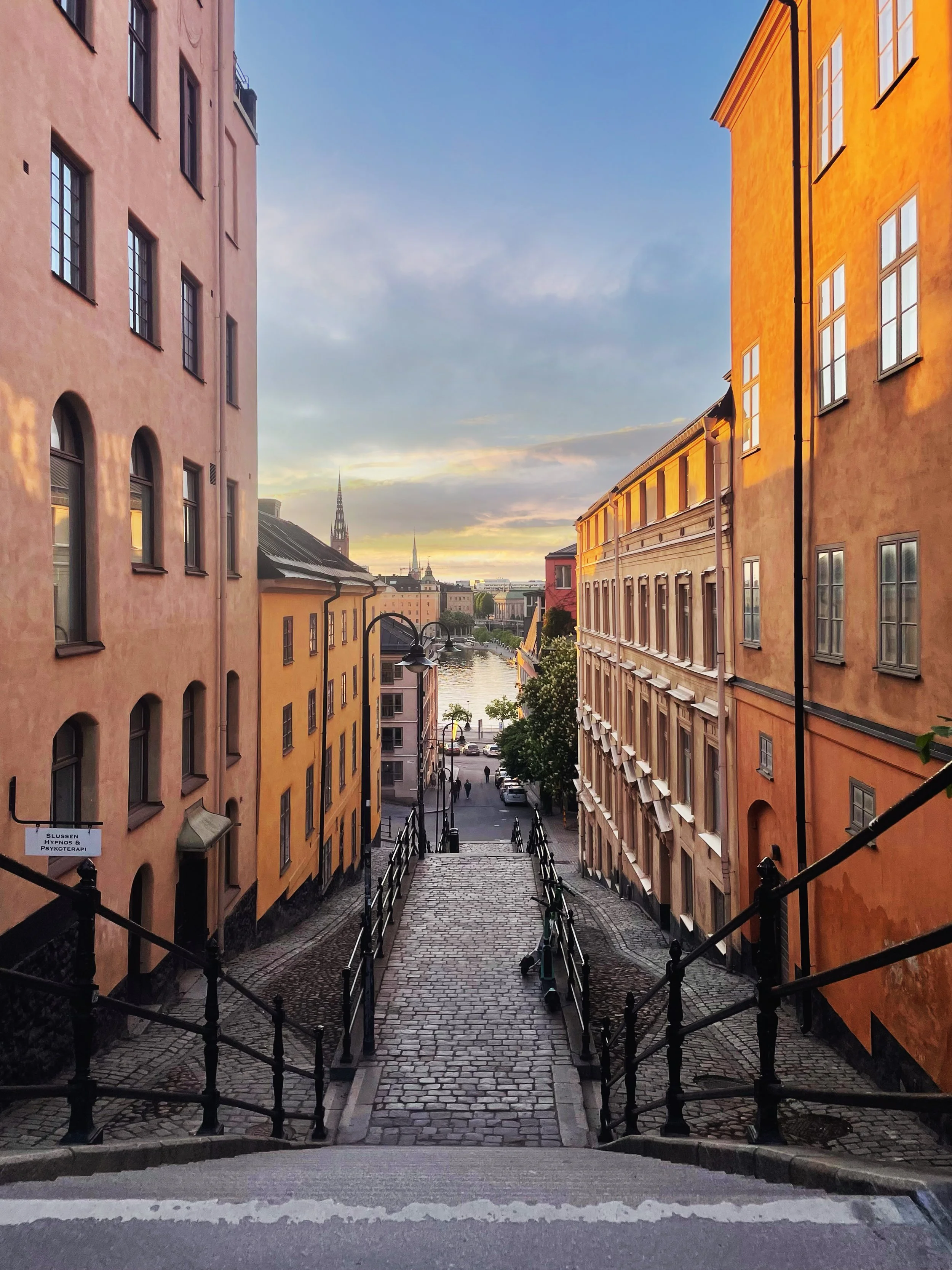 A cobblestone street with black iron railings leading down towards a river, lined with colorful buildings on both sides, under a partly cloudy sky at sunset.