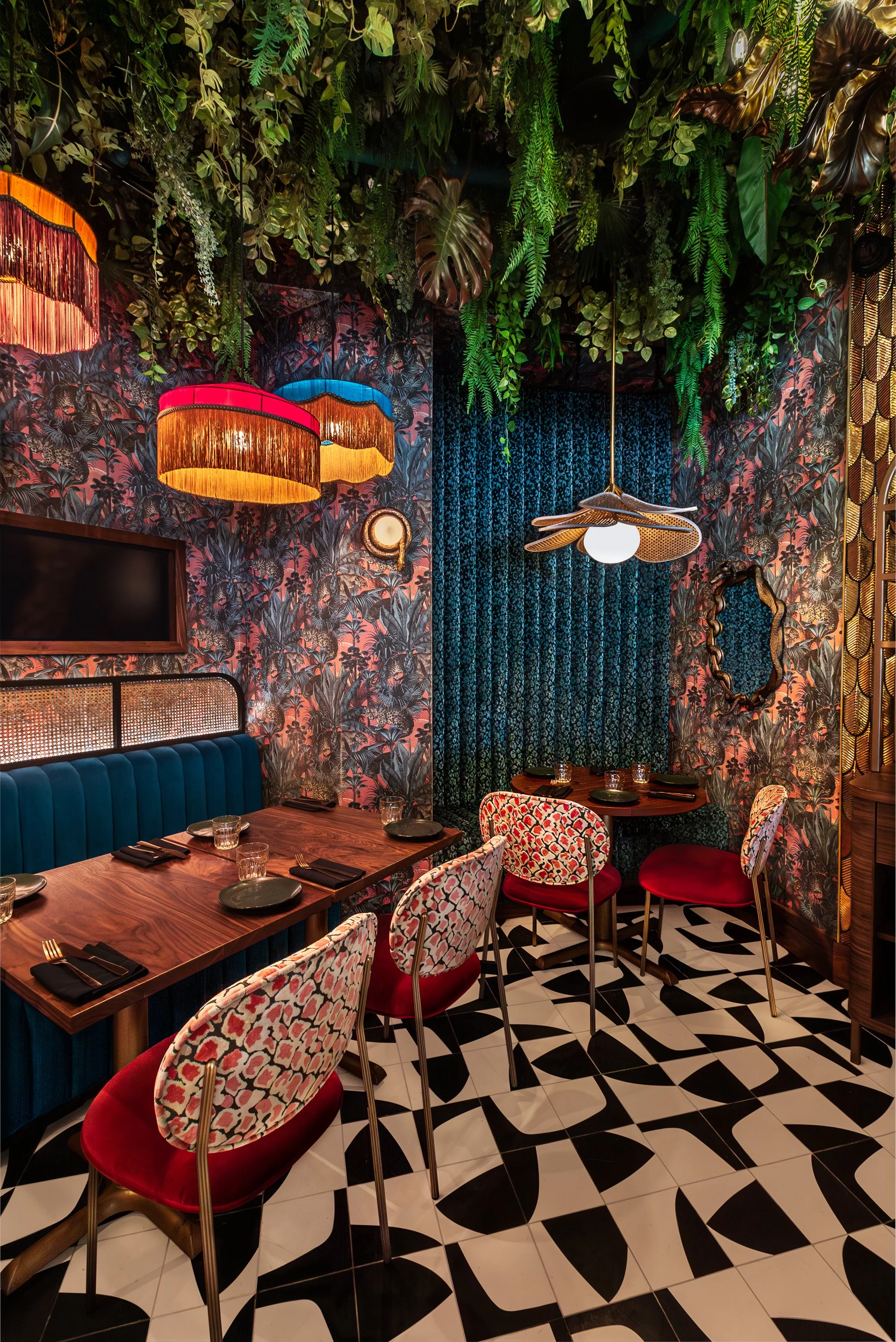 A vibrant restaurant interior with tropical foliage ceiling decor, colorful hanging lamps, patterned wallpaper, a flat-screen TV, a blue upholstered bench, a wooden table set with black plates and glasses, and a mix of patterned chairs on a black and