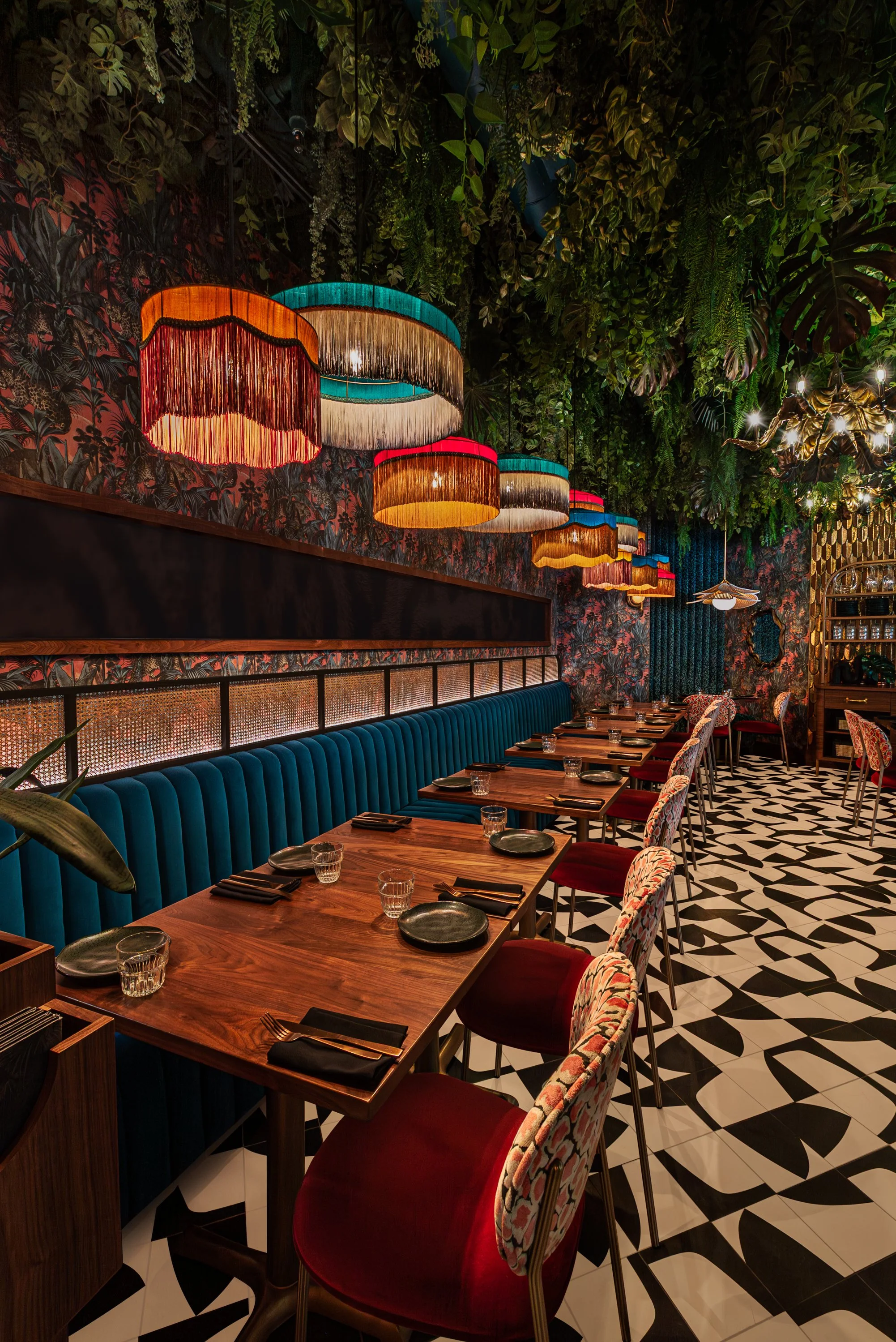 A warmly lit restaurant interior with colorful hanging lamps, a long blue velvet banquette, patterned chairs with red cushions, and black-and-white geometric floor tiles. Lush greenery decorates the ceiling and walls.