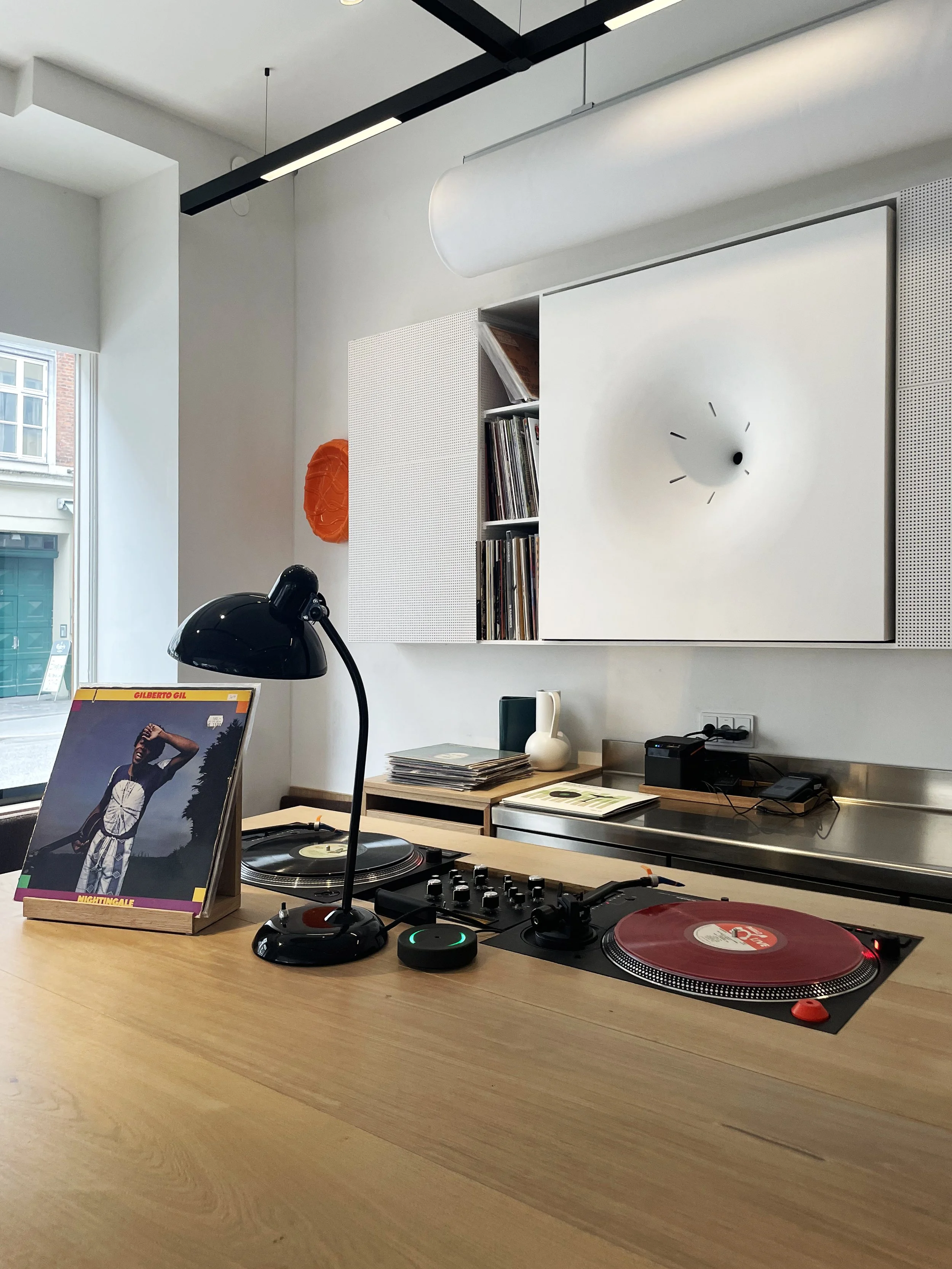A modern interior with a wooden table featuring DJ equipment, vinyl records, a black lamp, and a window in the background.