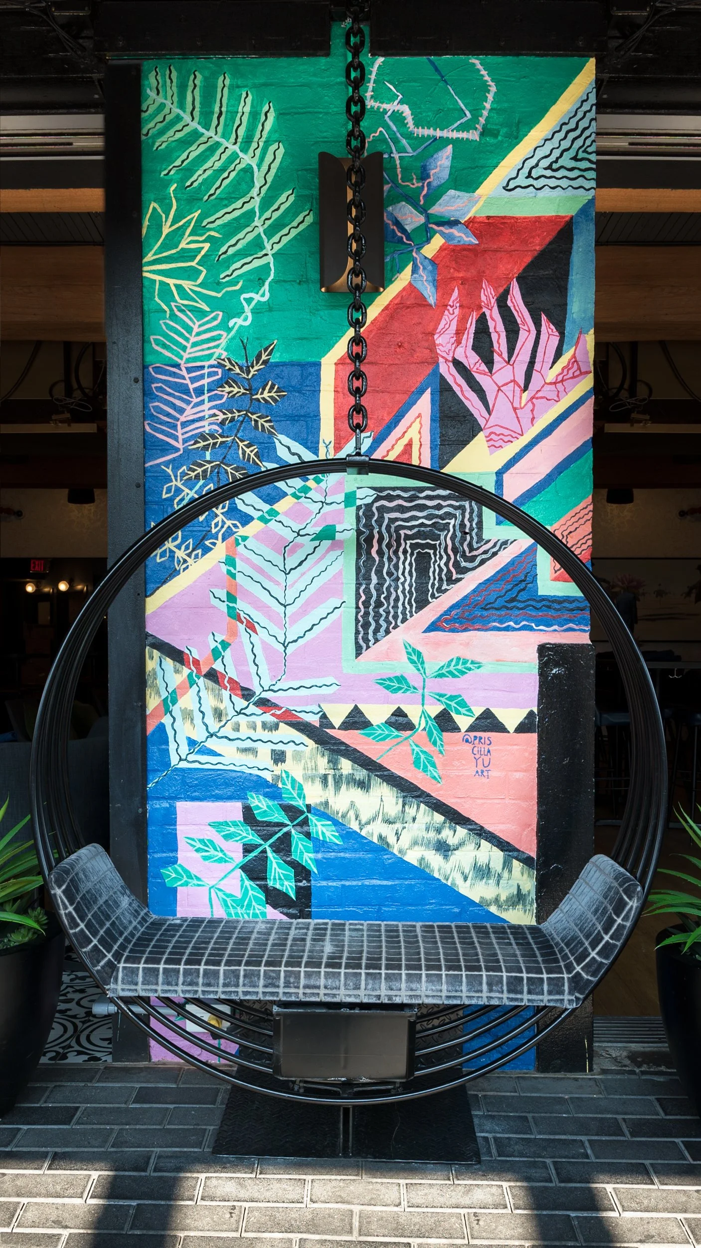 Colorful mural painted on a brick wall featuring abstract leaves and geometric shapes. There is a hanging circular chair with a padded seat in front of the mural, and two potted plants on either side.
