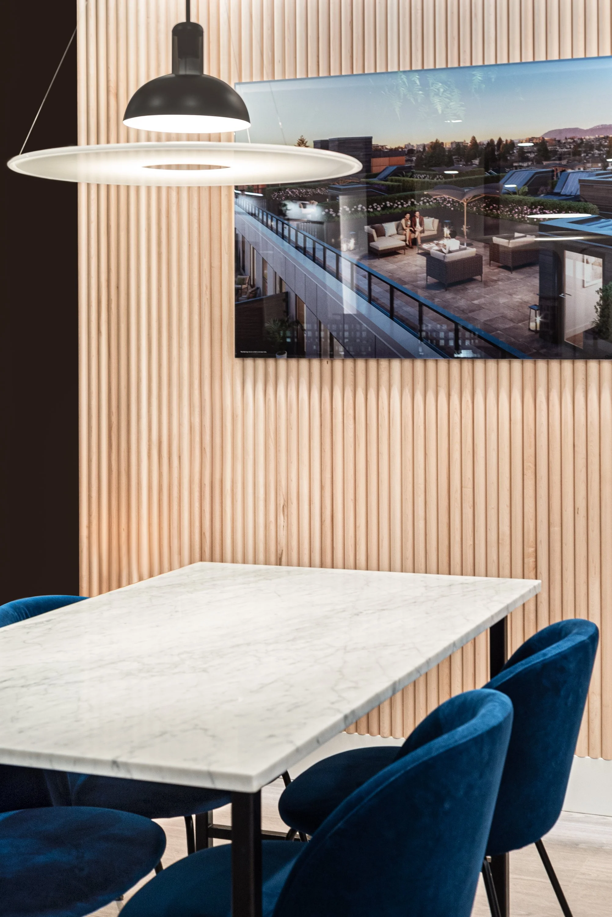 Interior of a modern dining area with a white marble table, blue velvet chairs, a wooden slat wall, a large hanging pendant light, and a TV screen showing an outdoor rooftop terrace with seating and city view.