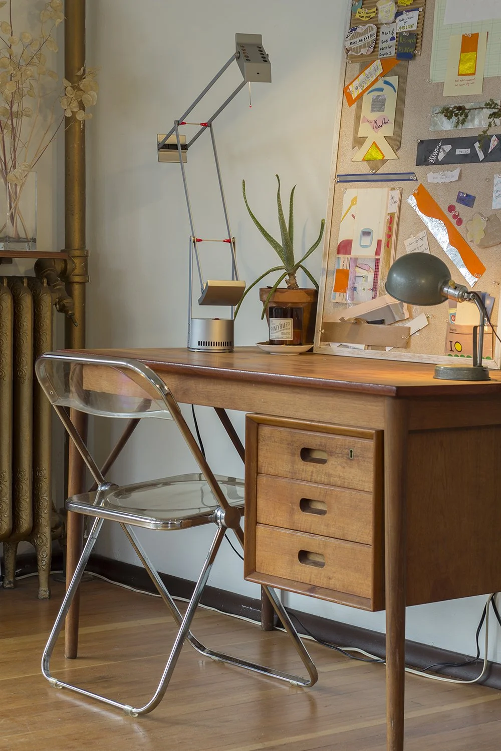 A wooden desk with three drawers on the right side, a modern clear acrylic and metal chair, a green desk lamp, a potted cactus, and a bulletin board with colorful papers and notes in the background.