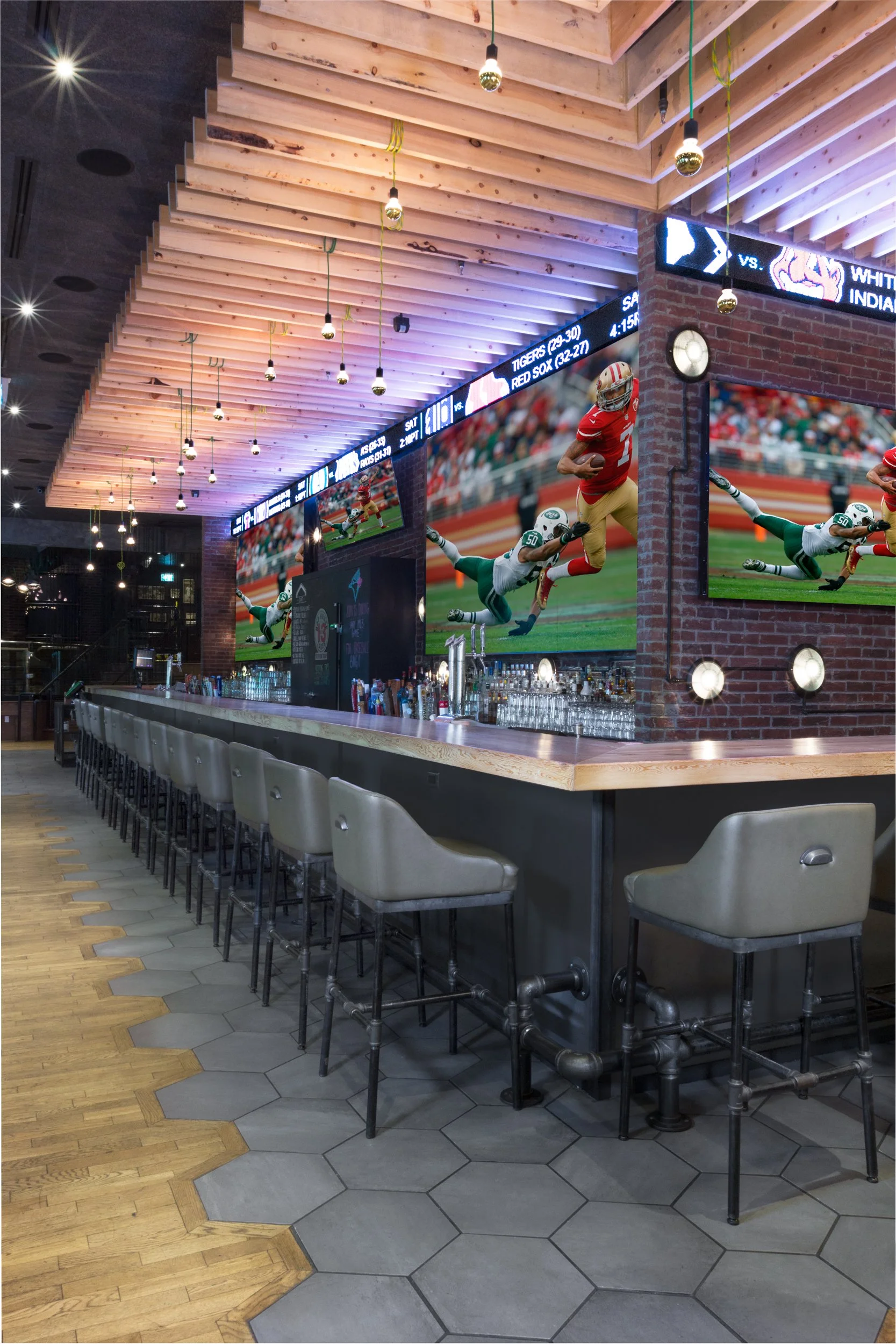 Modern sports bar with a long bar counter, barstools, and multiple large TV screens showing a football game. The ceiling features wooden slats and hanging light fixtures, with a brick wall background.