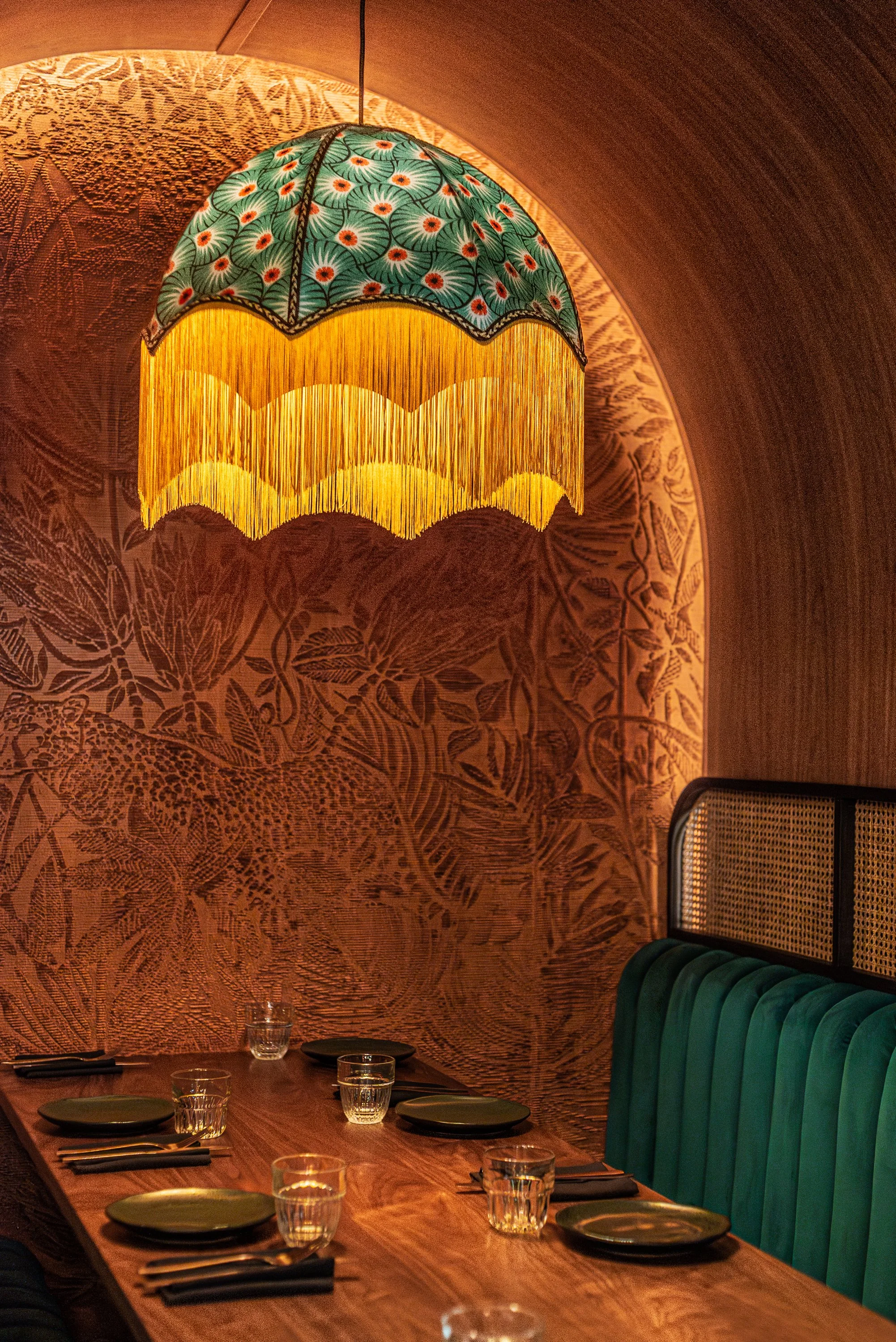 A wooden dining table set with six black plates, glasses of water, and black napkins, with a teal upholstered banquette against a wall with botanical patterns. A hanging lampshade with floral and fringe details illuminates the scene.