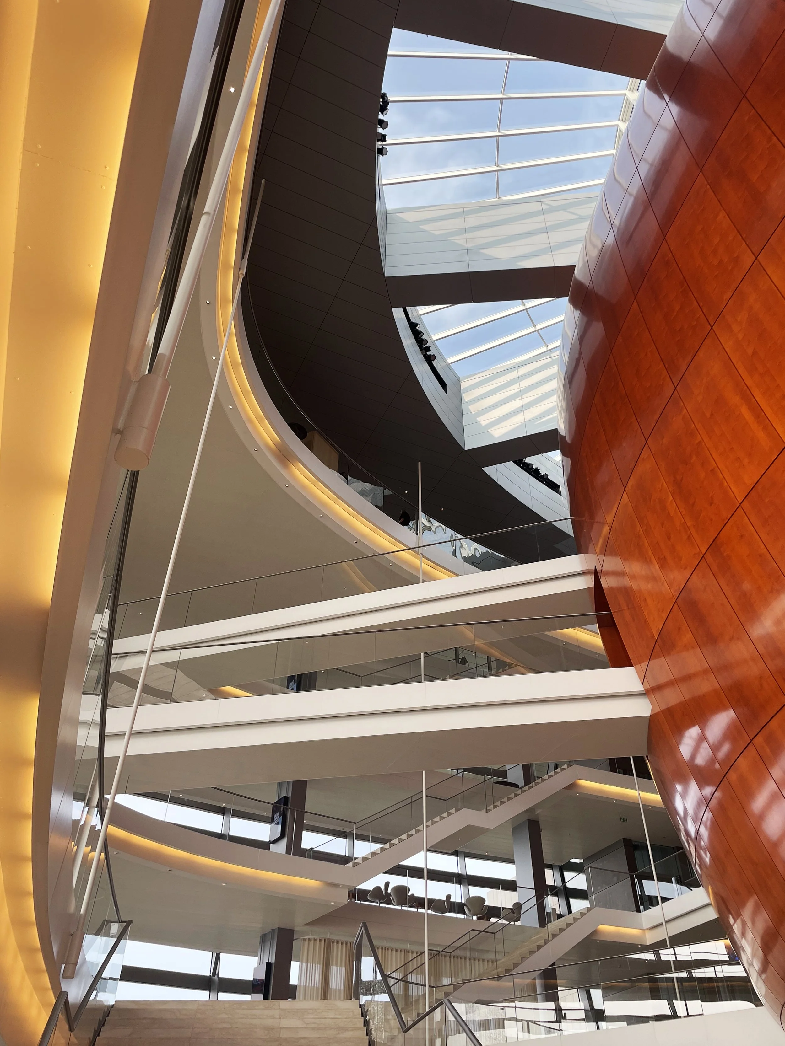 Interior view of a modern multi-level building with glass ceilings, curved wooden wall, and white walkways with glass railings.
