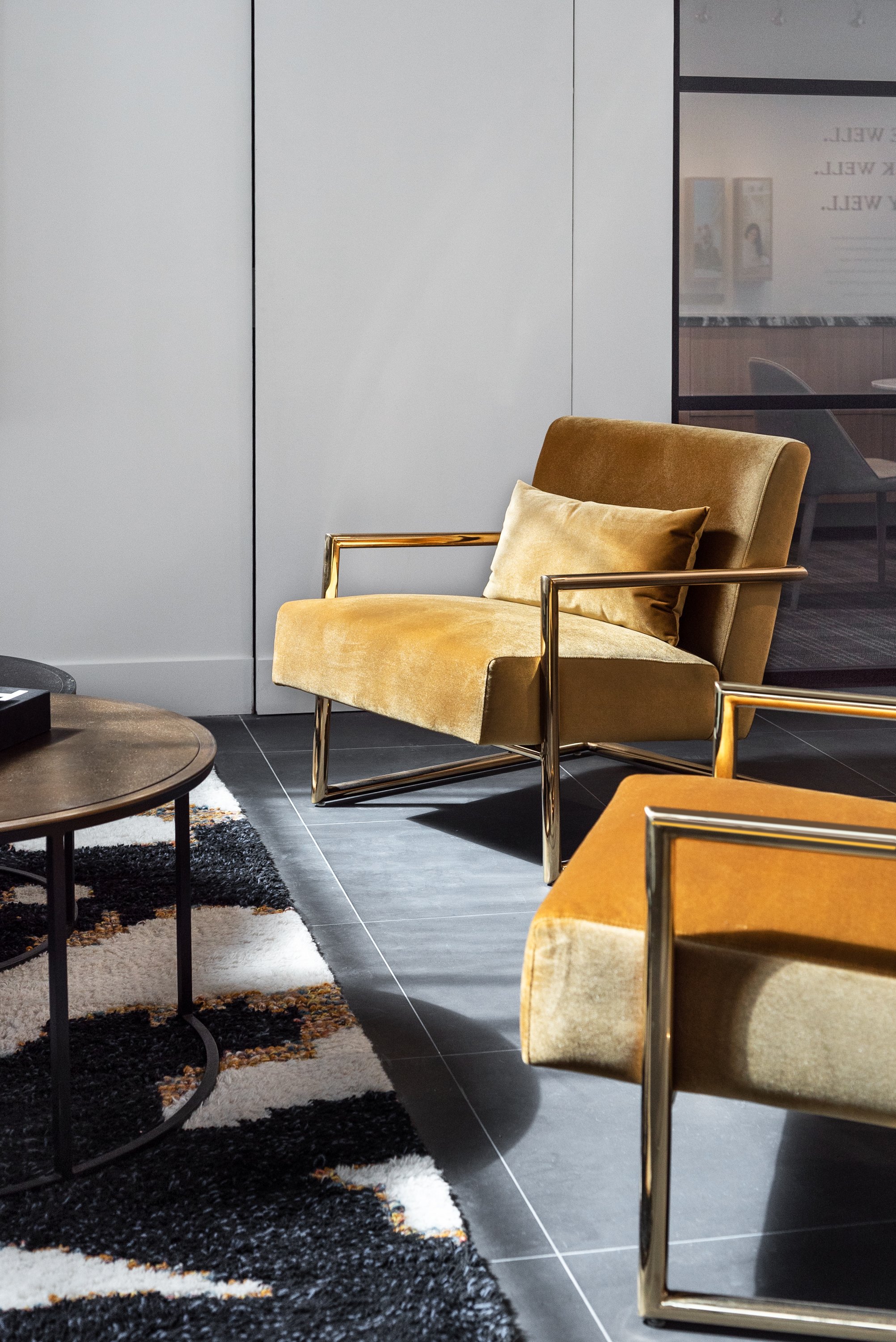 Modern interior with yellow velvet armchairs with gold metal frames, a black coffee table, a rug with black, white, and brown patterns, and a glass wall showing a conference room.