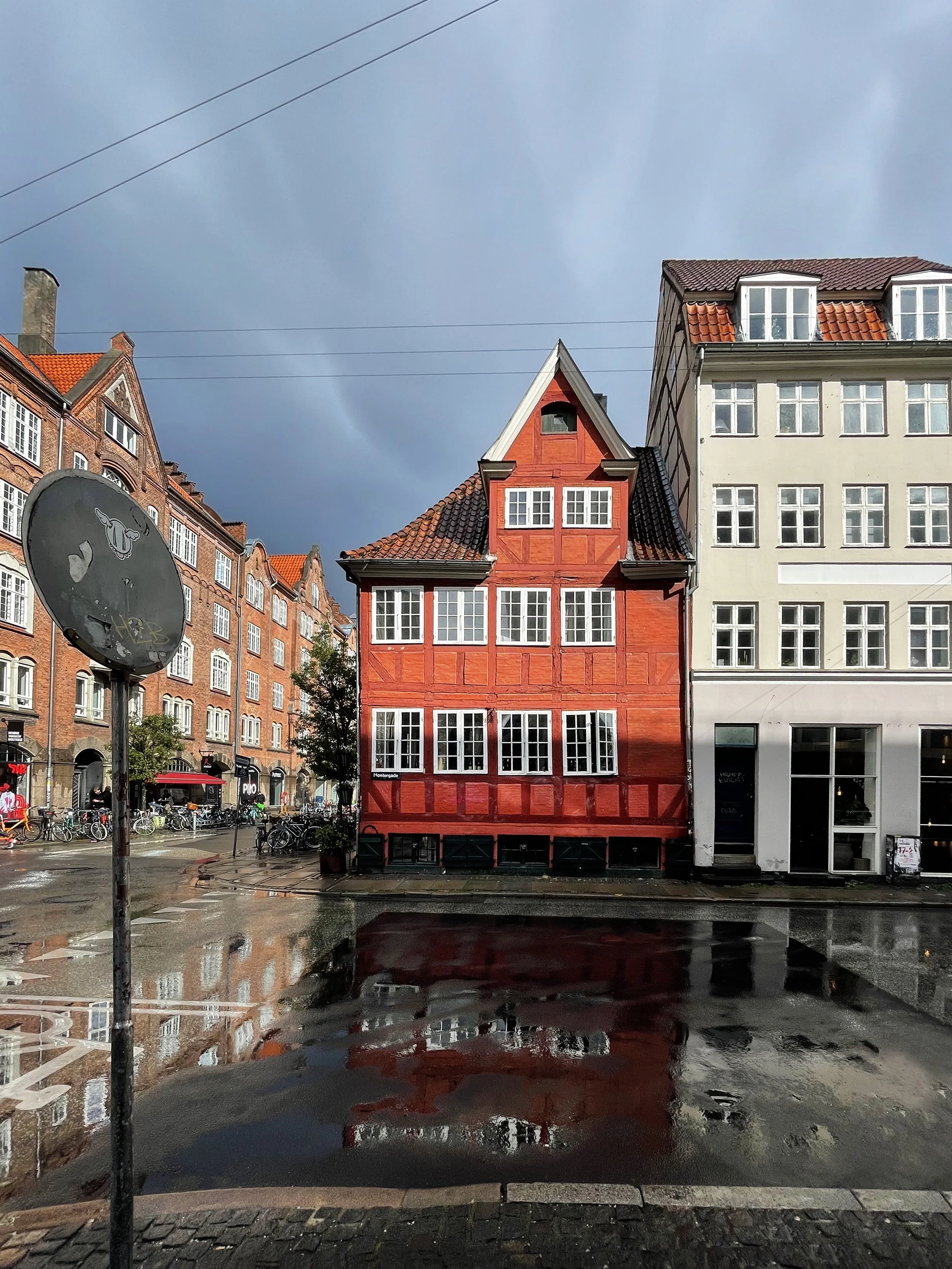 Colorful buildings on a city street after rain, with a large black bicycle sign and puddles reflecting the buildings.
