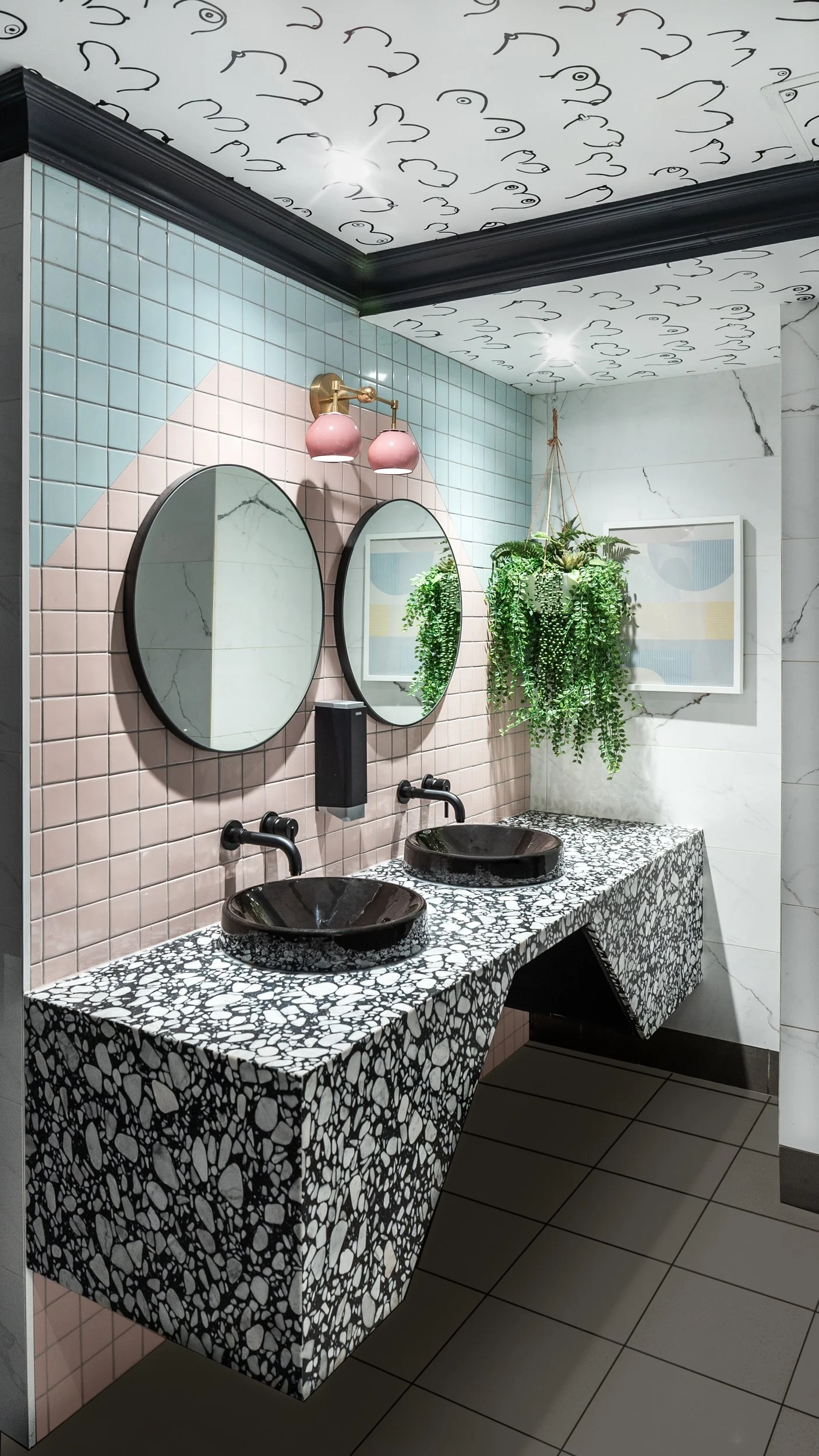 A modern bathroom with a terrazzo countertop with two black vessel sinks and black wall-mounted faucets. Two round mirrors are hung on a pink tiled wall, above which is a pink light fixture. A large green hanging plant is on the white marble wall wit
