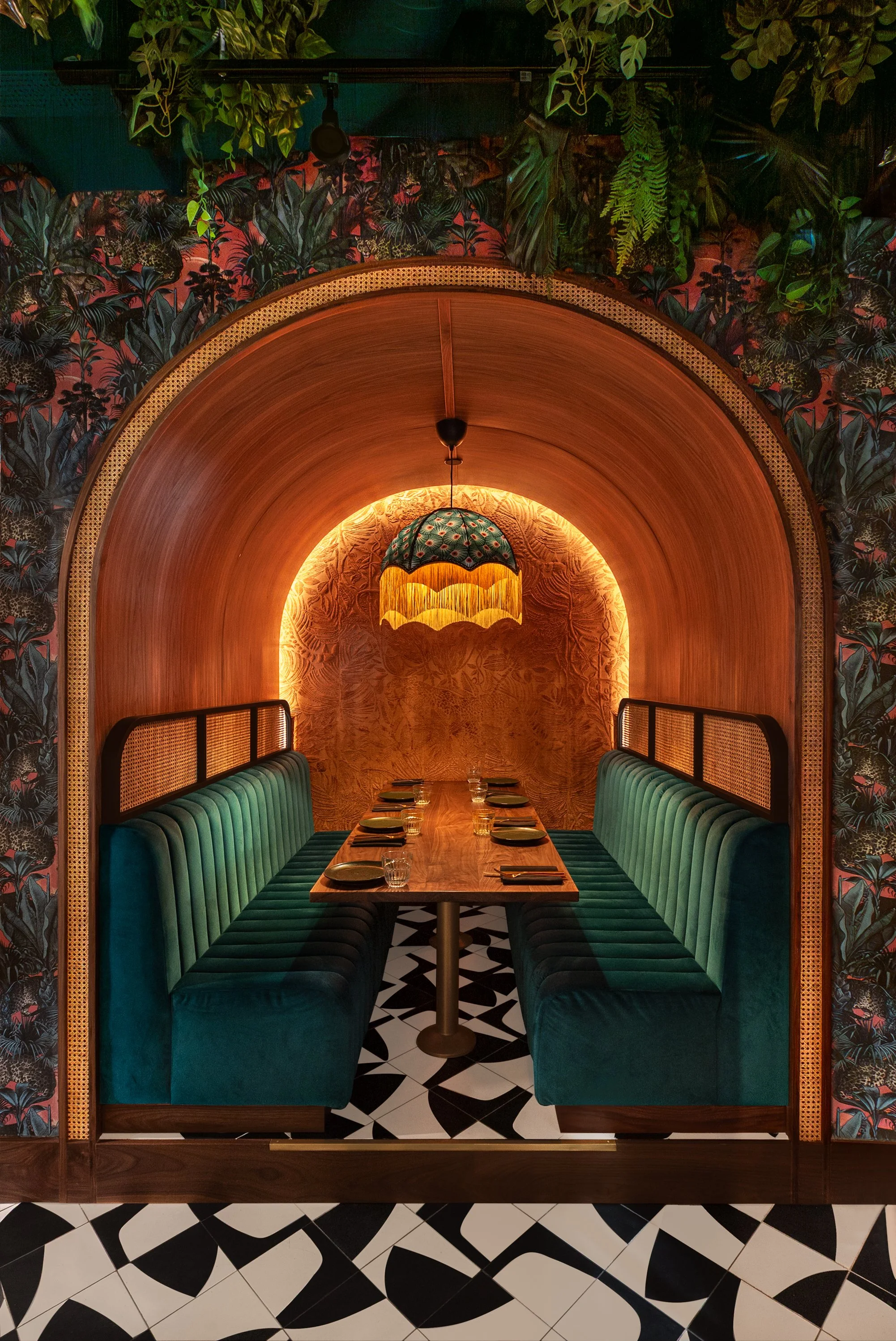 An intimate dining nook with teal velvet benches, a wooden table set with plates and glasses, framed by a curved wooden arch, greenery and tropical wallpaper on the walls, a decorative hanging lamp, and patterned black and white floor tiles.