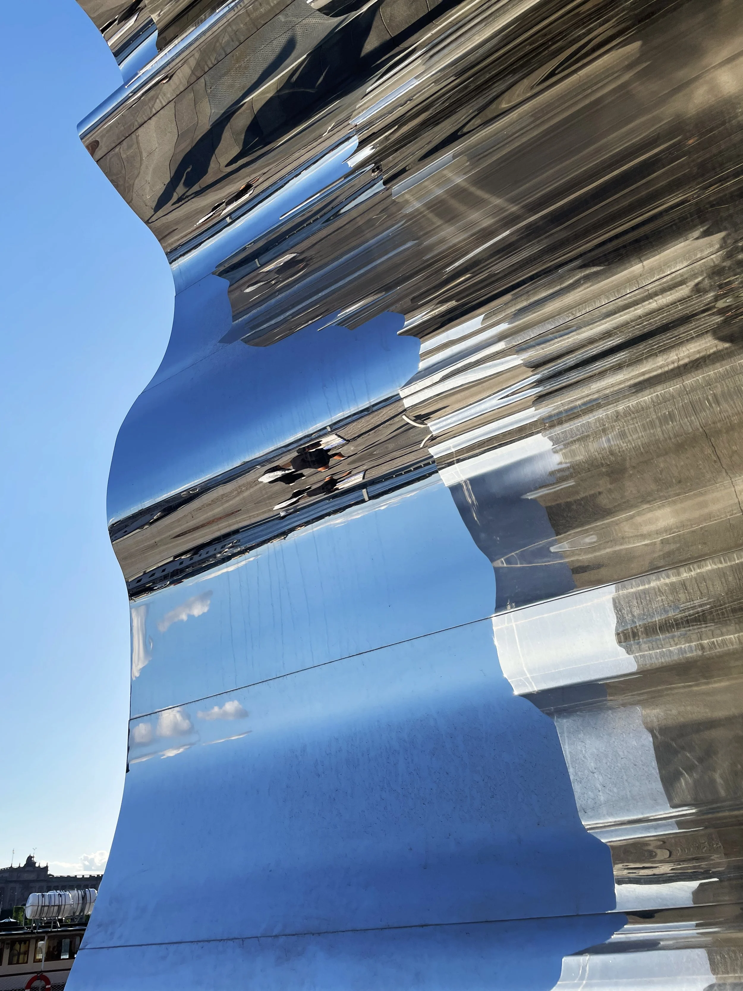 Close-up of a shiny, reflective metal sculpture with curving and jagged edges, reflecting the blue sky and surroundings.