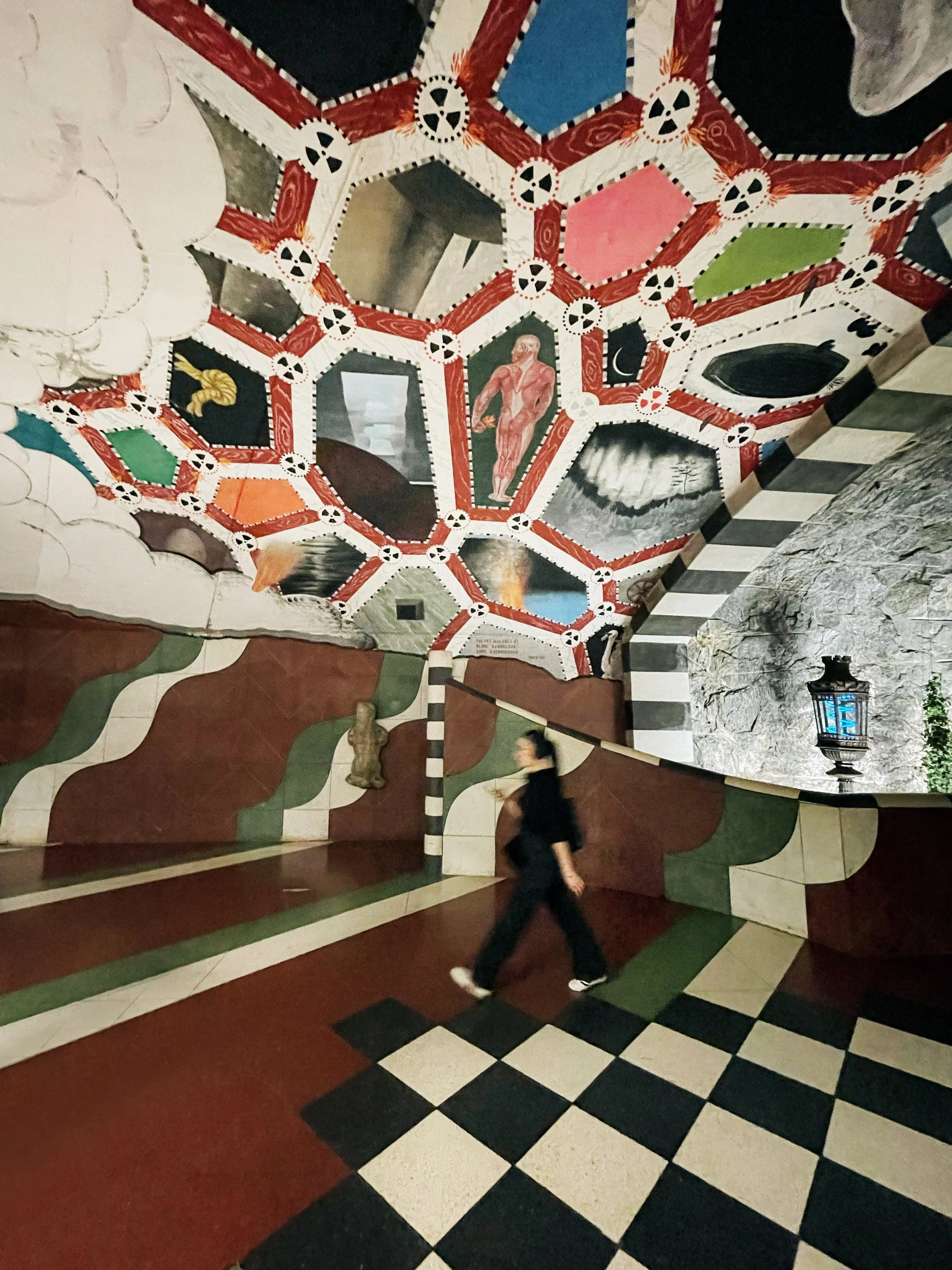 A person walking through a colorful, surreal room with painted walls and ceiling featuring geometric patterns, abstract images, and figures, including a human figure and celestial elements, with a black-and-white checkered floor.