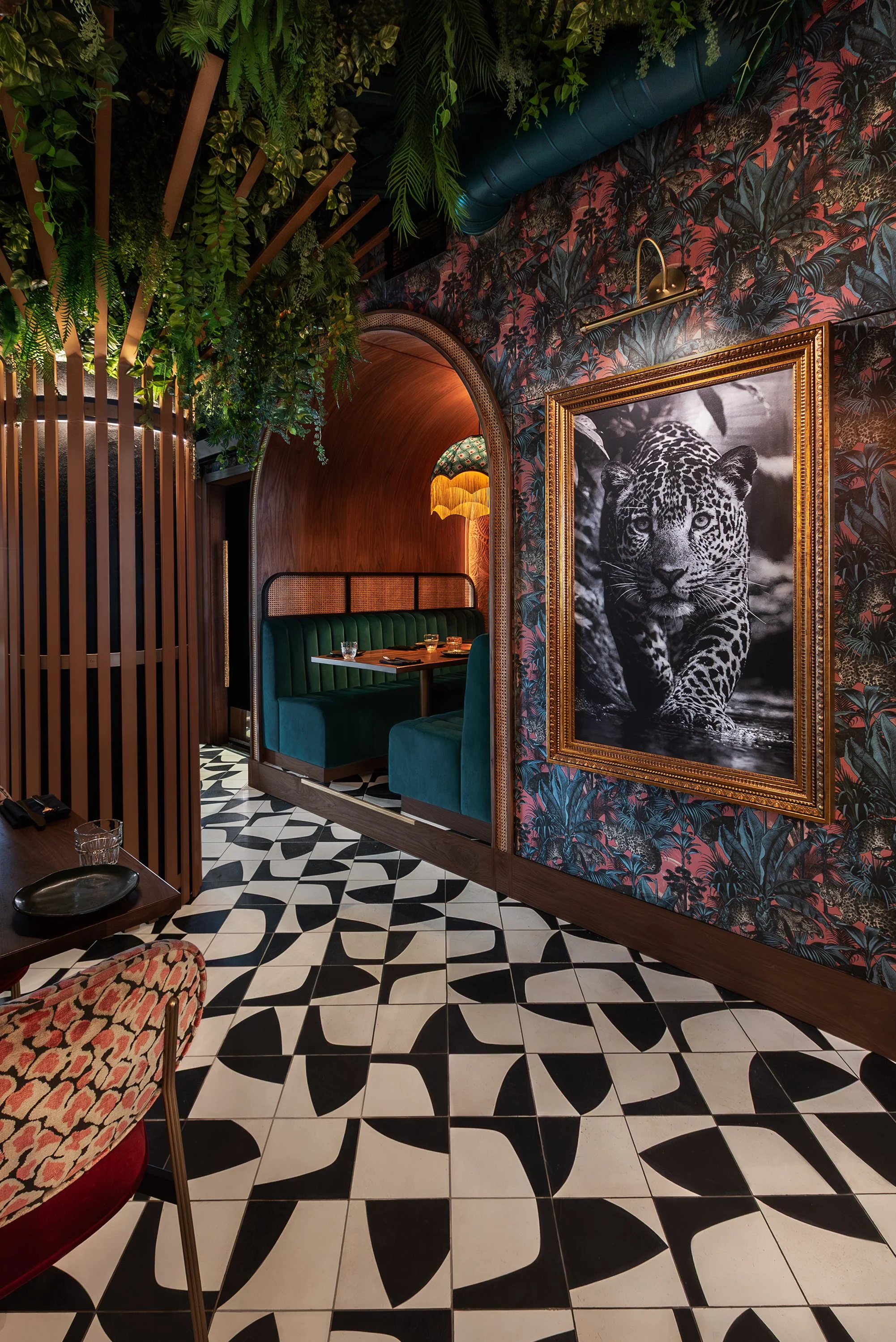 Interior of a restaurant with black and white patterned floor tiles, a large leopard photograph in a gold frame on a tropical-themed wallpapered wall, a green booth dining area with teal velvet seating, hanging light fixtures, and lush greenery overh