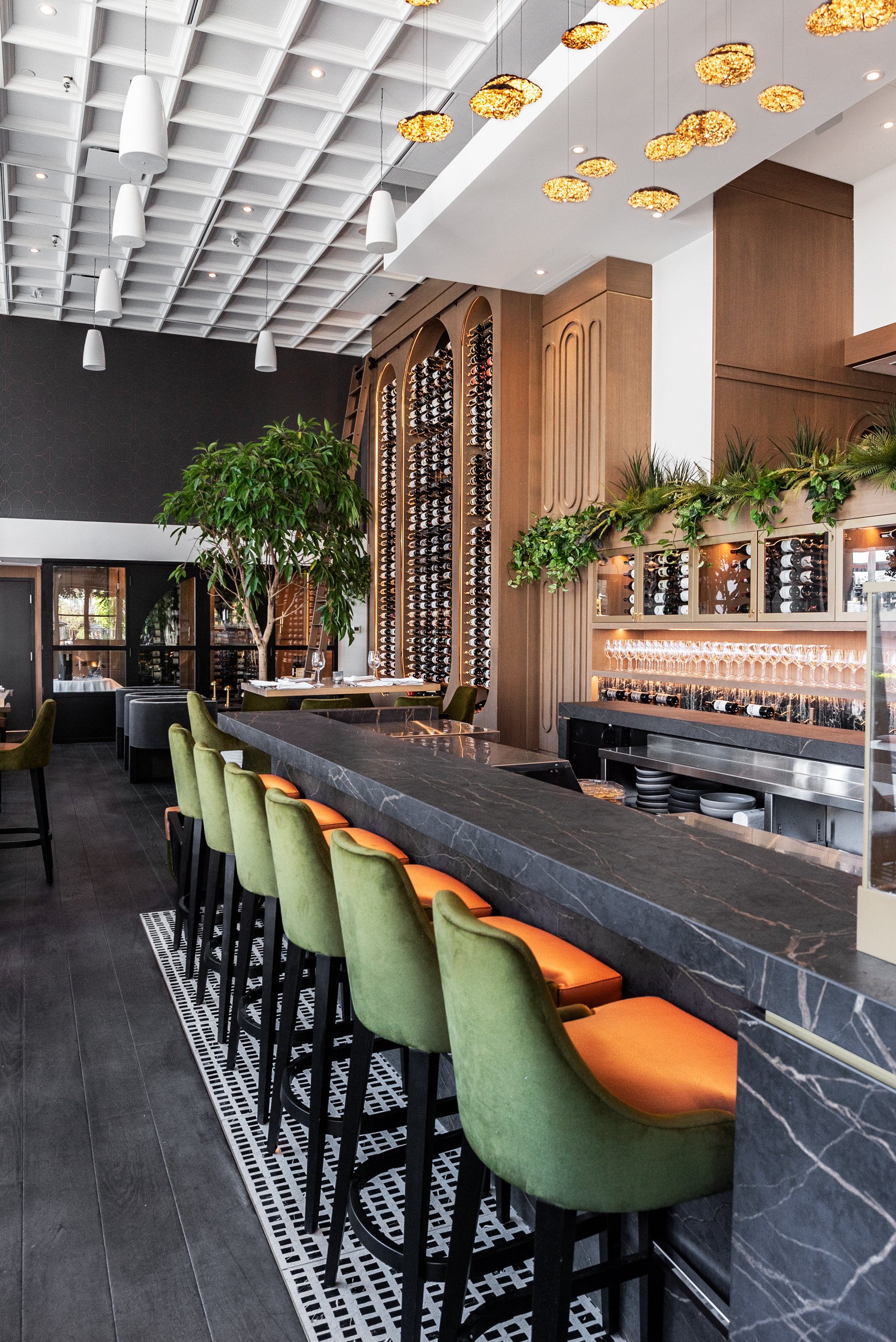 A modern restaurant interior featuring a black marble bar with green and orange upholstered bar stools. Behind the bar, there are wine racks built into wooden paneling, decorative greenery, and a glass case. The ceiling has a geometric pattern with h