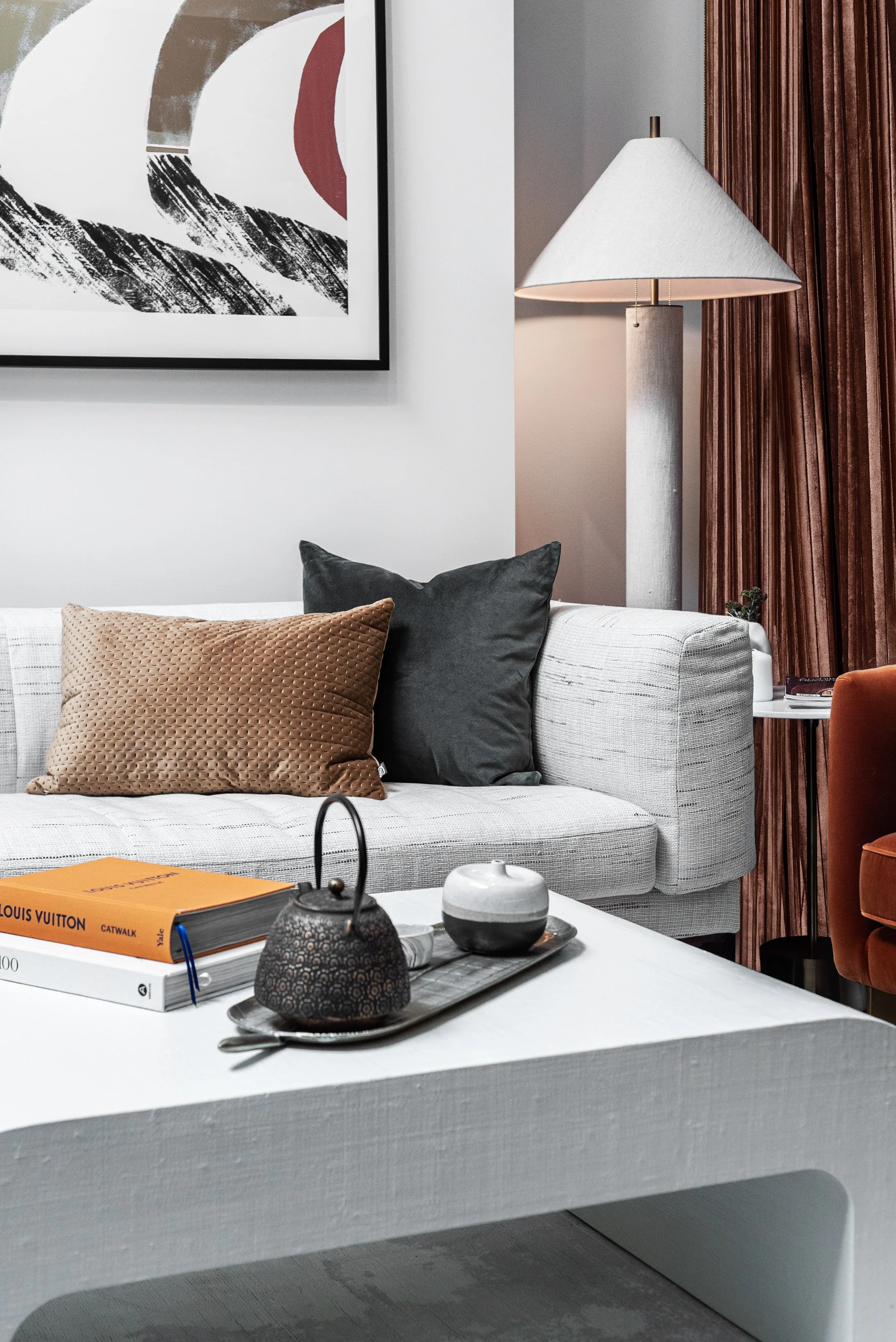 Interior of a modern living room with a white sofa, black and beige pillows, a white coffee table with decorative items, a gray and white abstract painting, a tall white floor lamp, and brown curtains.