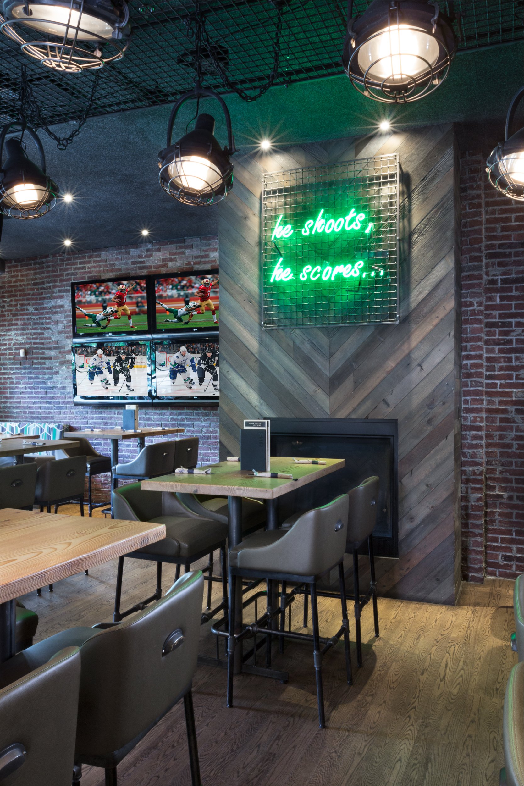 Interior of a sports bar with wooden and brick walls, multiple television screens displaying hockey games, a neon sign on the wall that reads 'he shoots, he scores,' hanging lighting fixtures, and wooden tables with chairs.
