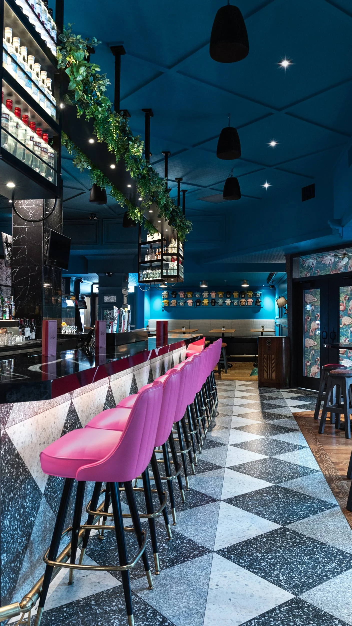 Interior of a bar with pink stools, a black marble counter, and a blue wall with decorative jars. Green plants hang from the ceiling, and there are black pendant lights, a checkered floor, and a wall with flamingo wallpaper.