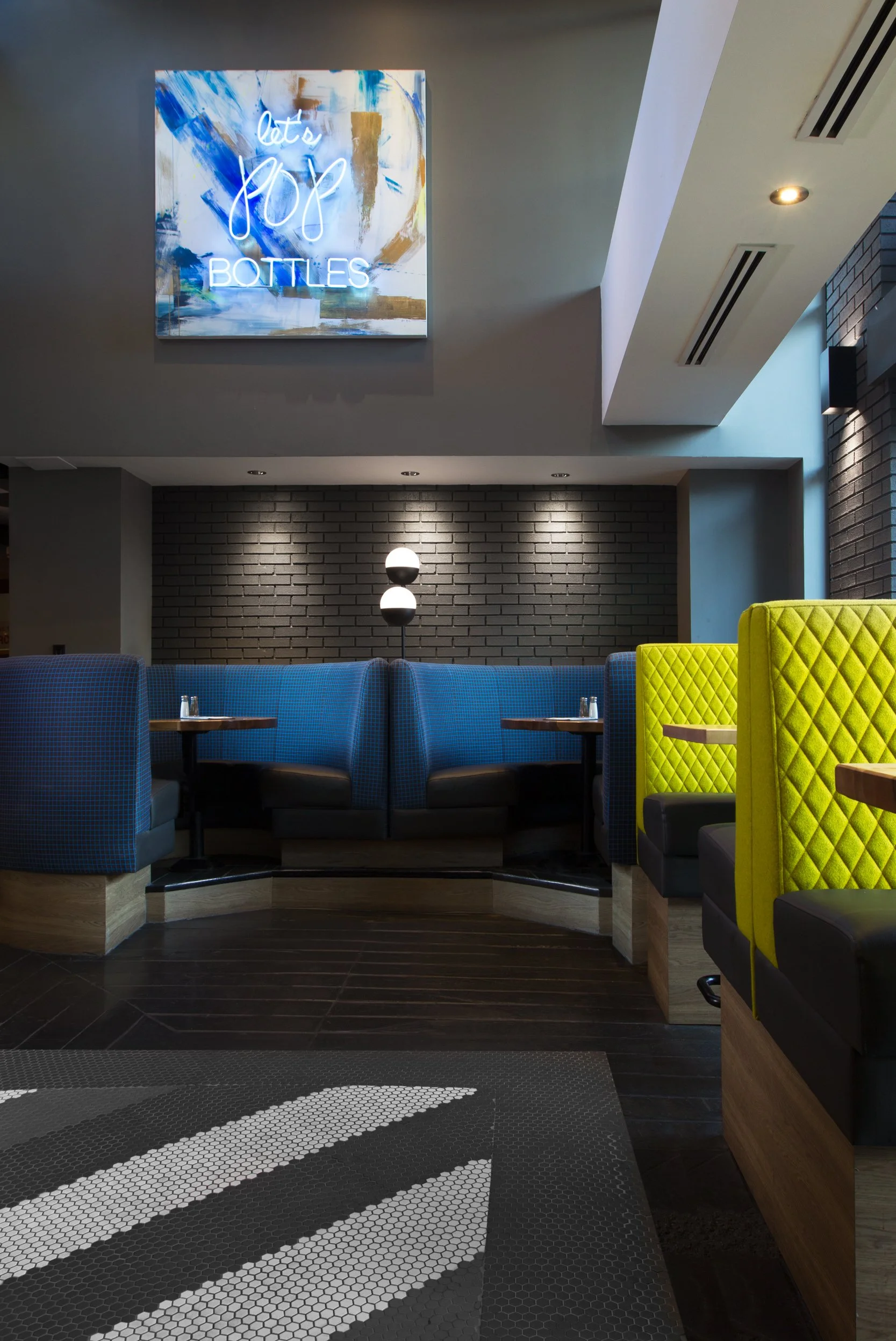 Interior of a restaurant with a neon sign that says "let's pop bottles," dark brick wall, booth seating with blue and yellow upholstery, and modern lighting.