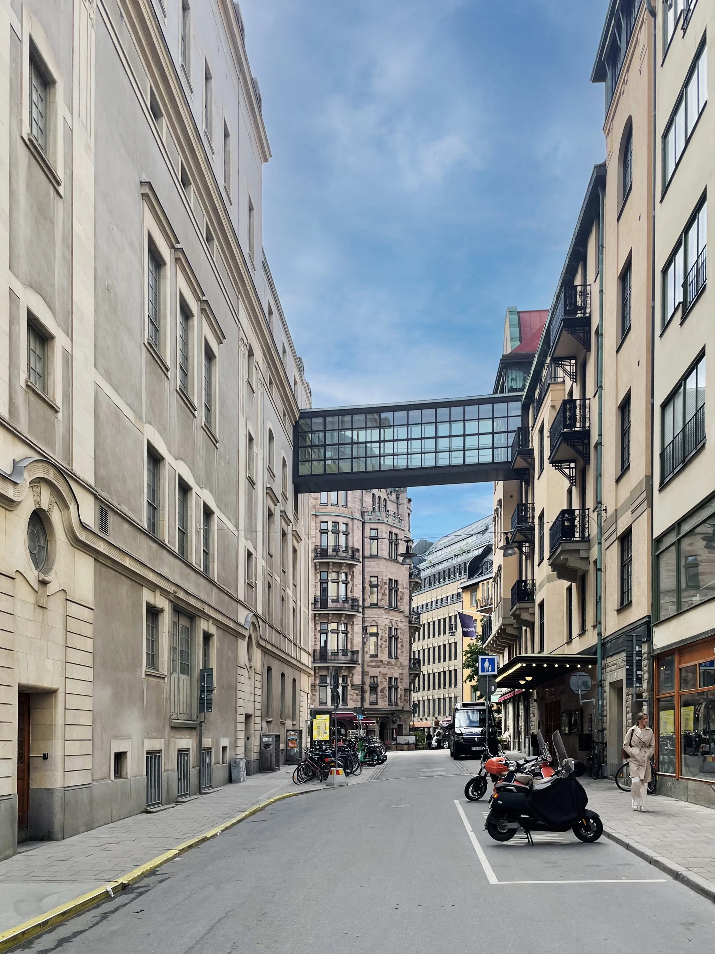 Urban street view with tall buildings, a sky bridge connecting buildings, parked motorcycles and bicycles, a pedestrian, and a delivery scooter.