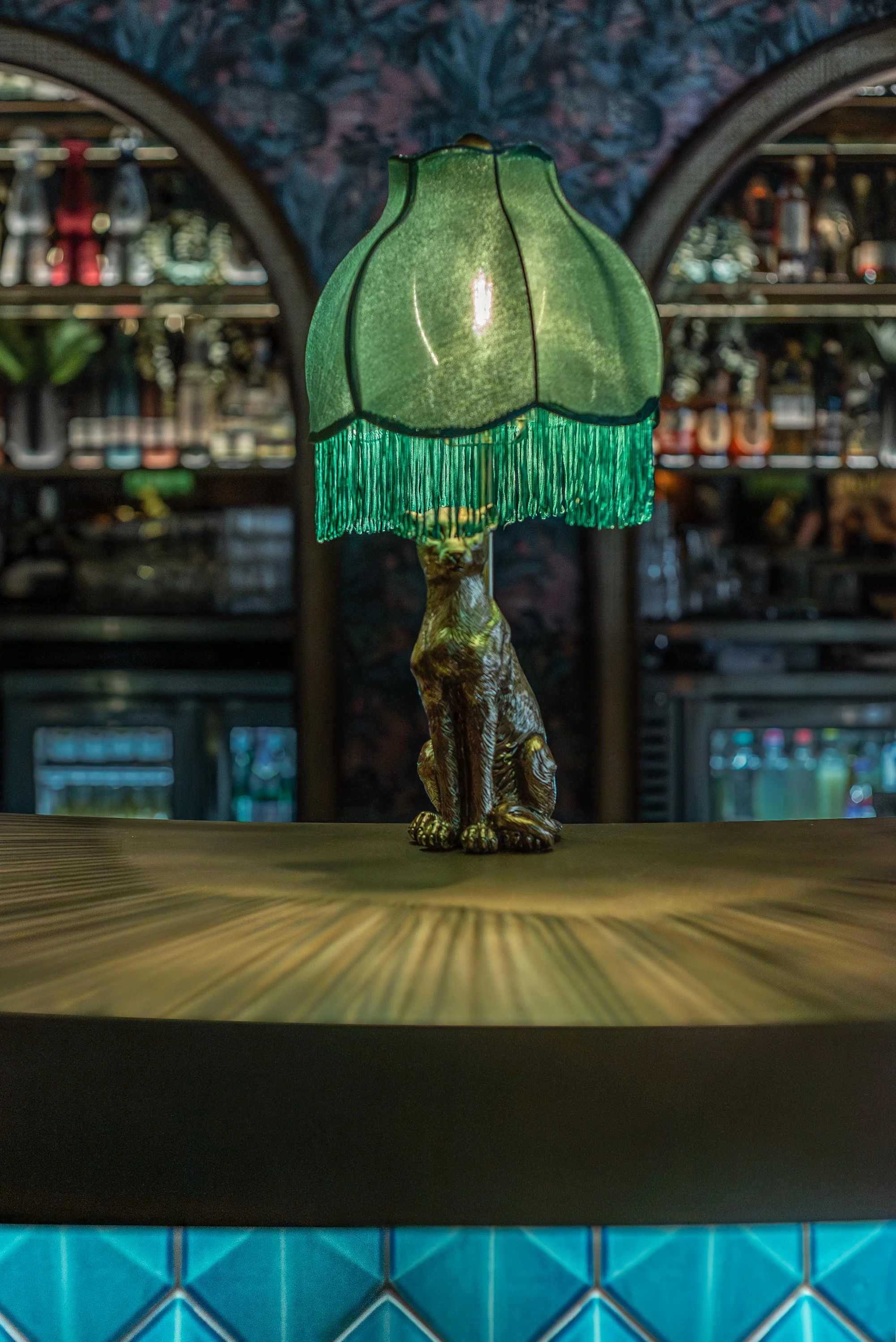 A decorative table lamp with a green fabric shade and fringe, featuring a cat-shaped base, placed on a wooden table in a dimly lit bar or restaurant.