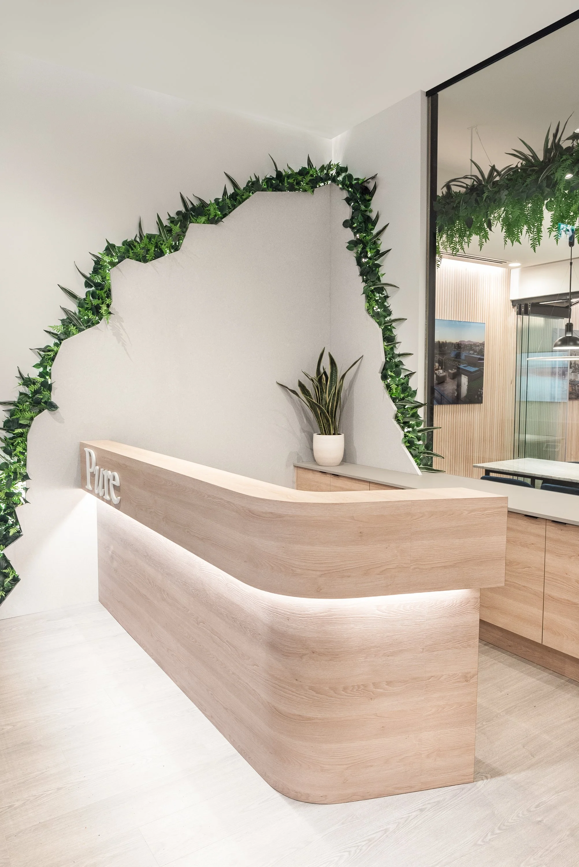 Modern reception desk with light wood finish and curved design, decorated with green plants and a sign that says "Pure", against a white wall with green leafy accents.