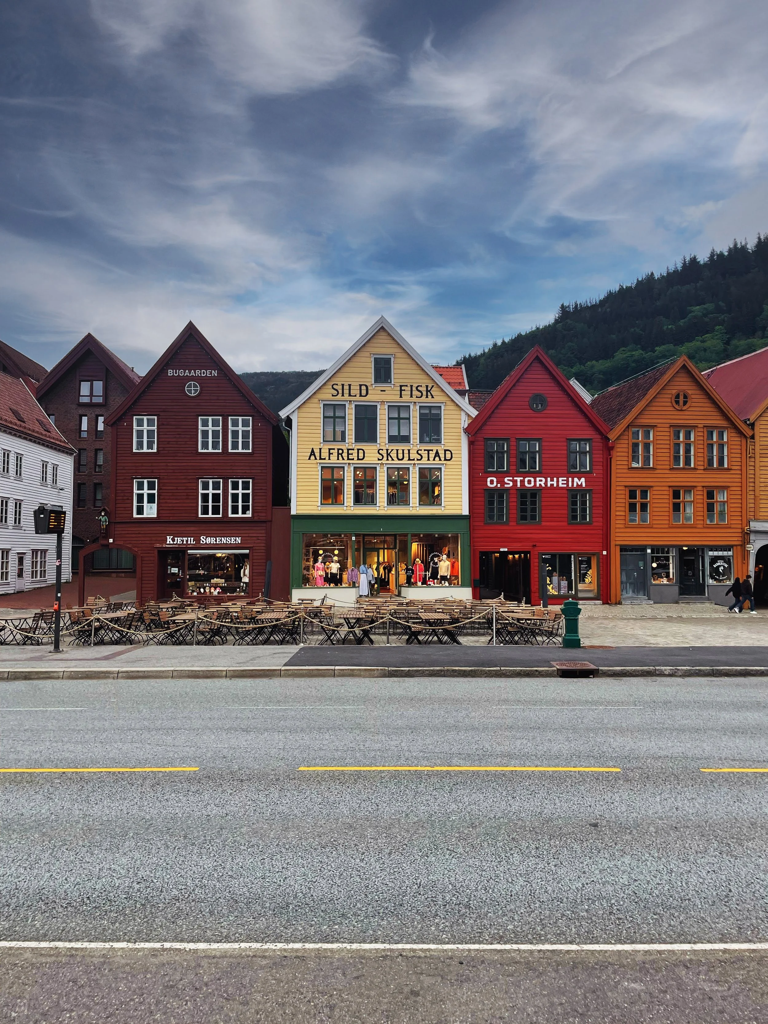 Colorful wooden buildings along a street, with outdoor seating in front, under a partly cloudy sky, in a scenic mountain town.