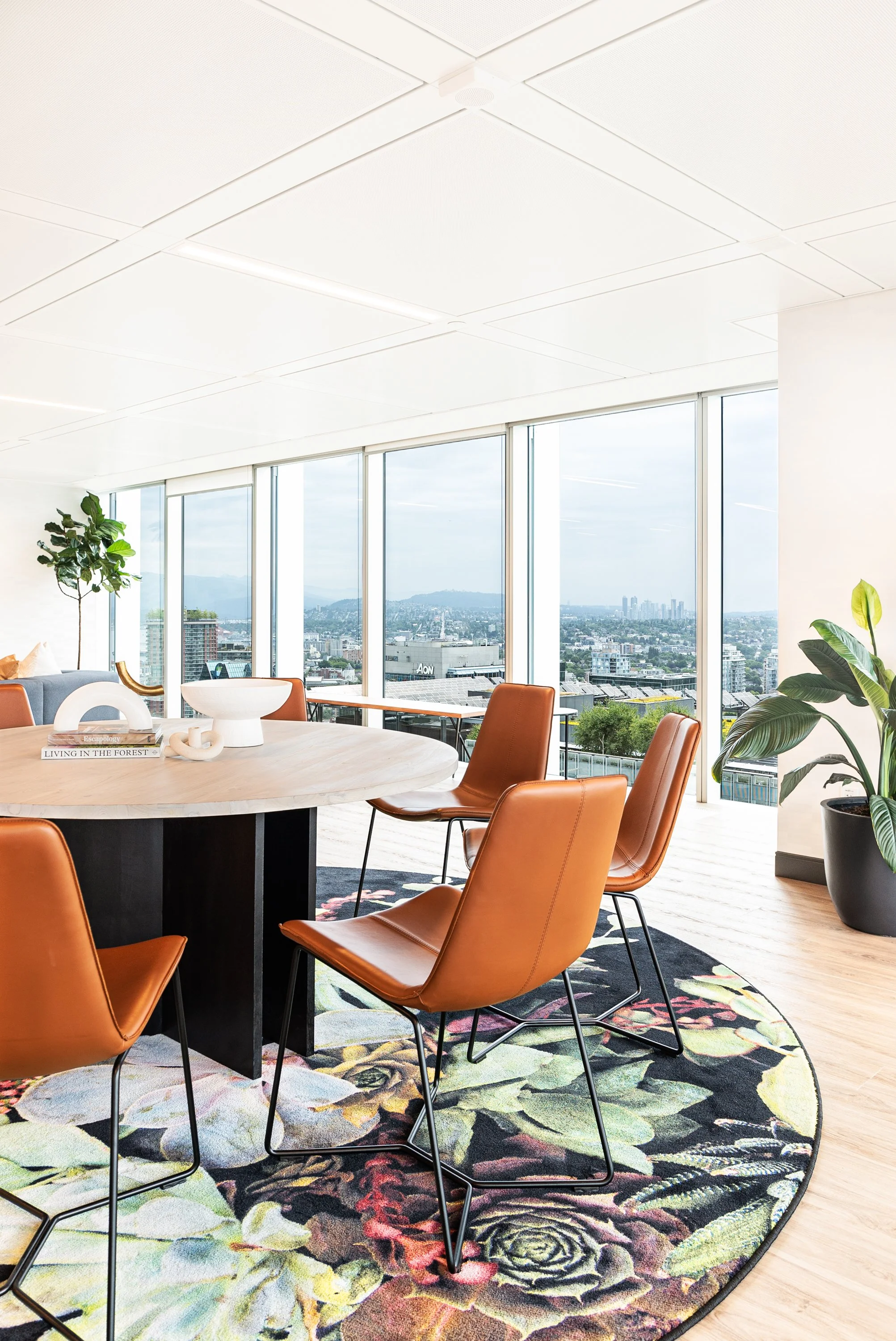 Modern conference room with a round table, brown chairs, large windows with cityscape view, indoor plants, and a floral rug.