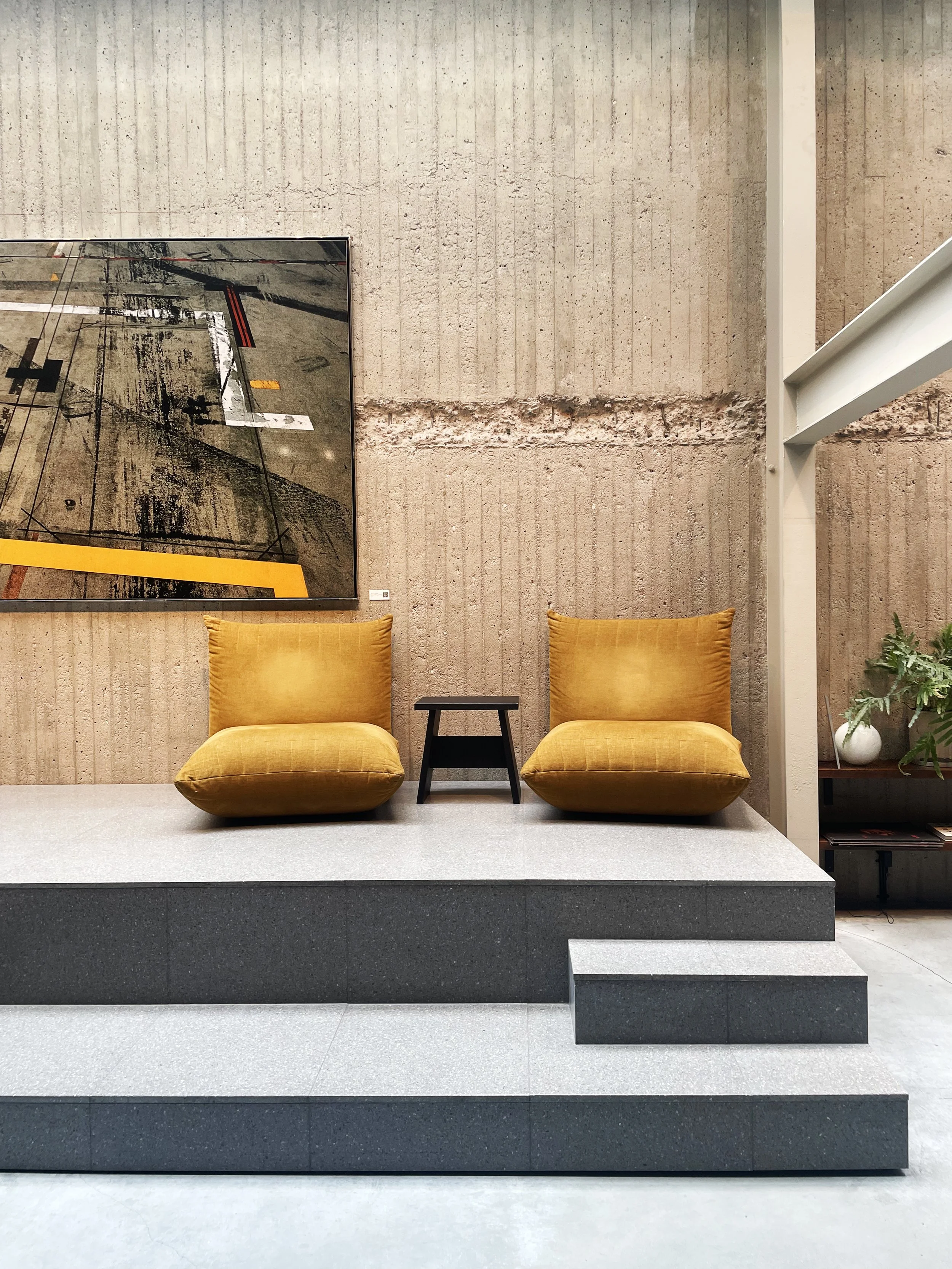 Two mustard yellow lounge chairs on a raised platform with a small black side table in between, against a textured beige wall with artwork, next to a plant on a shelf.