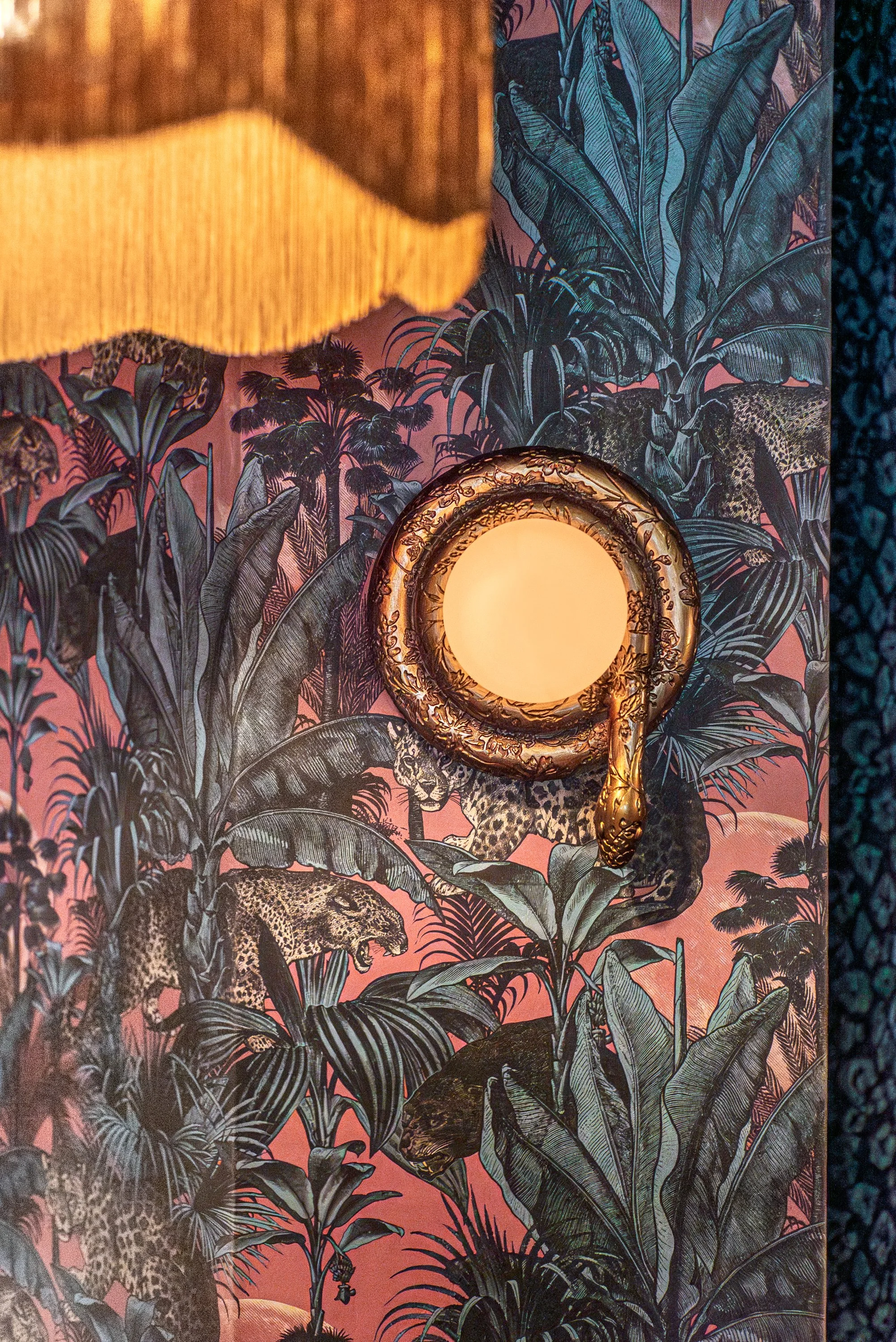 A wall covered in jungle-themed wallpaper featuring leopards and lush green plants, with a circular light fixture and a partial view of a yellow fabric lampshade.