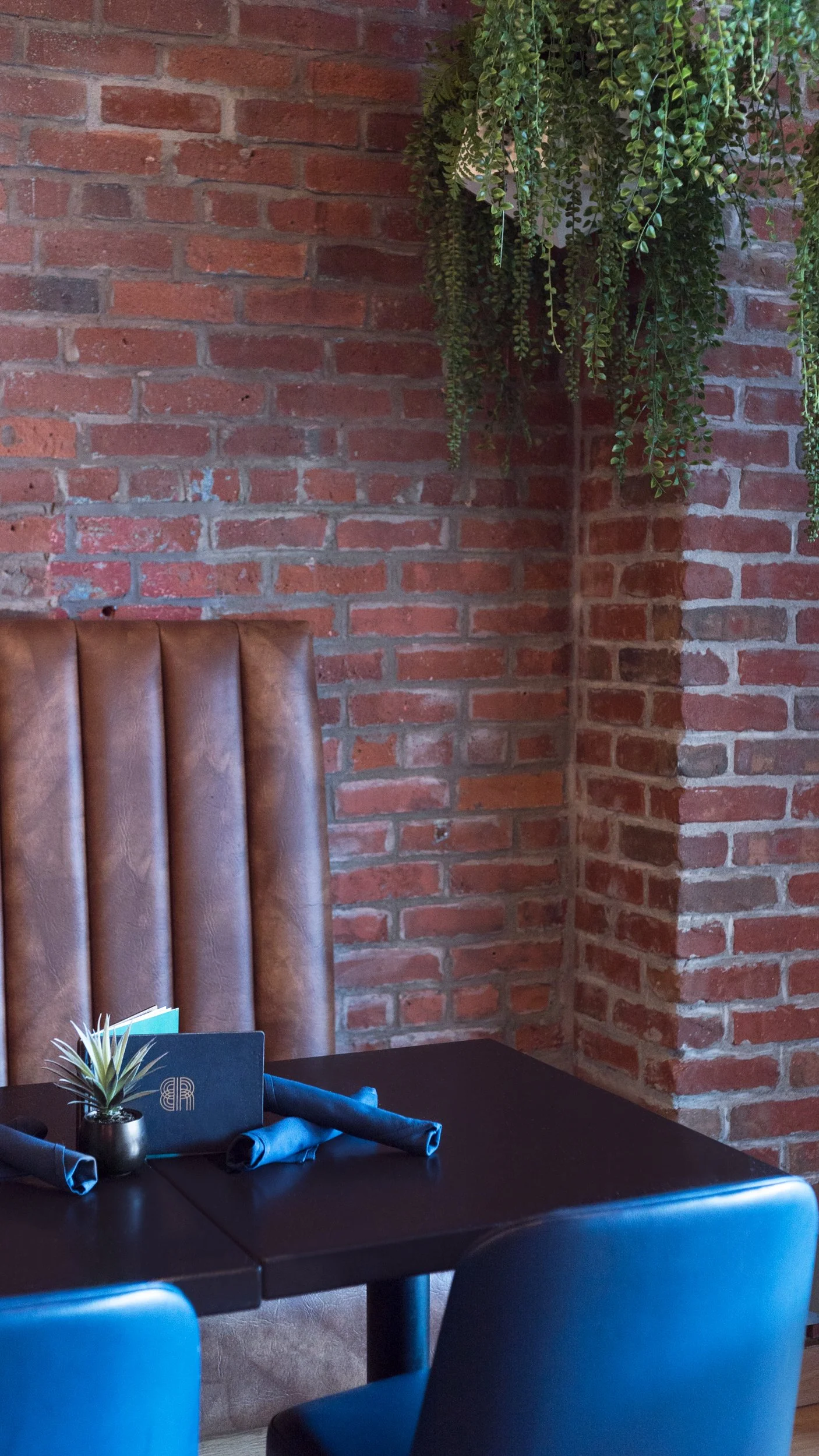 A cozy restaurant booth with a brown leather high-back seat, a dark wooden table, and black chairs. The table has a small potted plant, a menu, and rolled napkins. The background features an exposed red brick wall with hanging green plants.