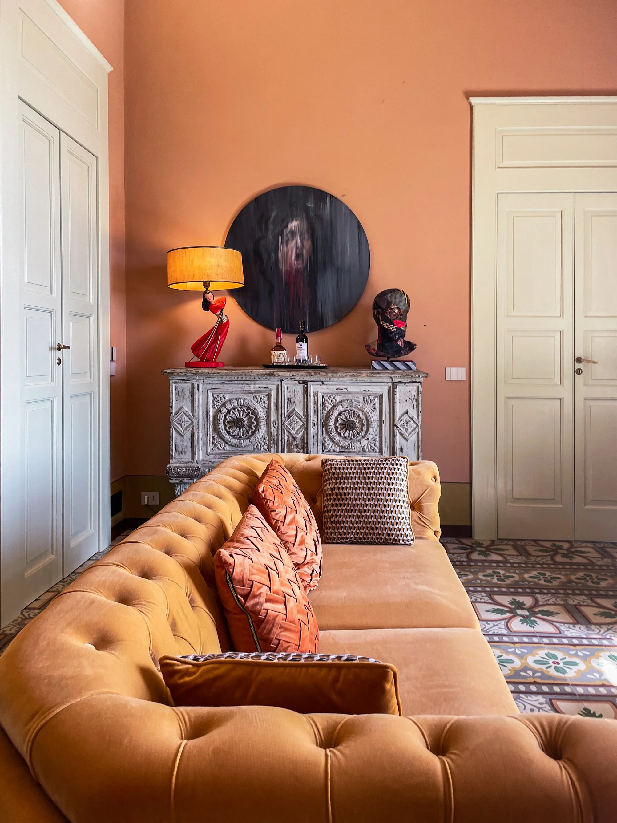 A hotel or lounge living room with a mustard yellow tufted sofa and four patterned throw pillows. In the background is a console table with decorative items, a large round mirror, a lamp, and a sculpture with a face mask. The walls are painted peach,