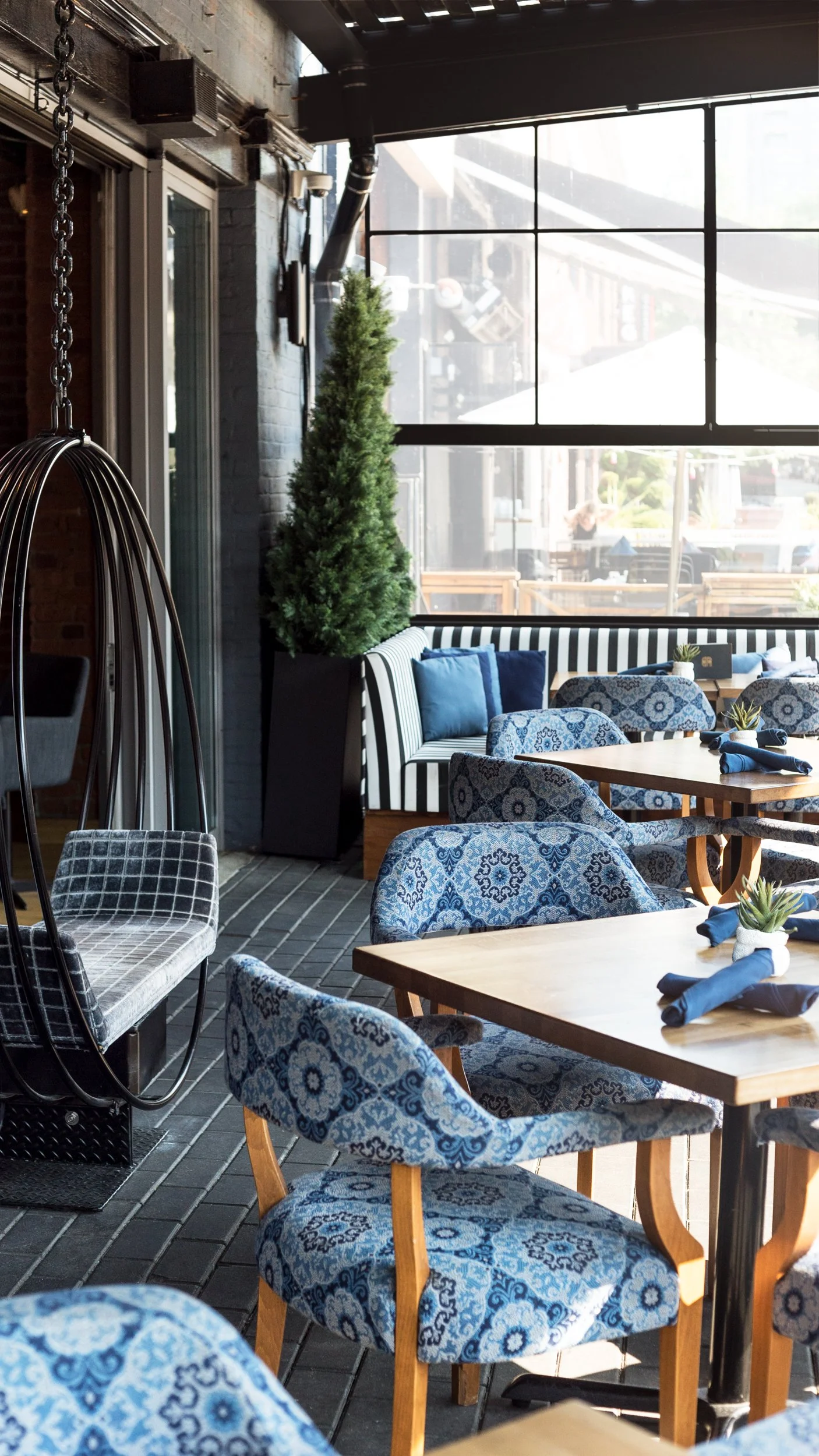 Interior of a modern restaurant with wooden tables, blue patterned chairs, striped booth seating, and large floor-to-ceiling windows allowing sunlight.