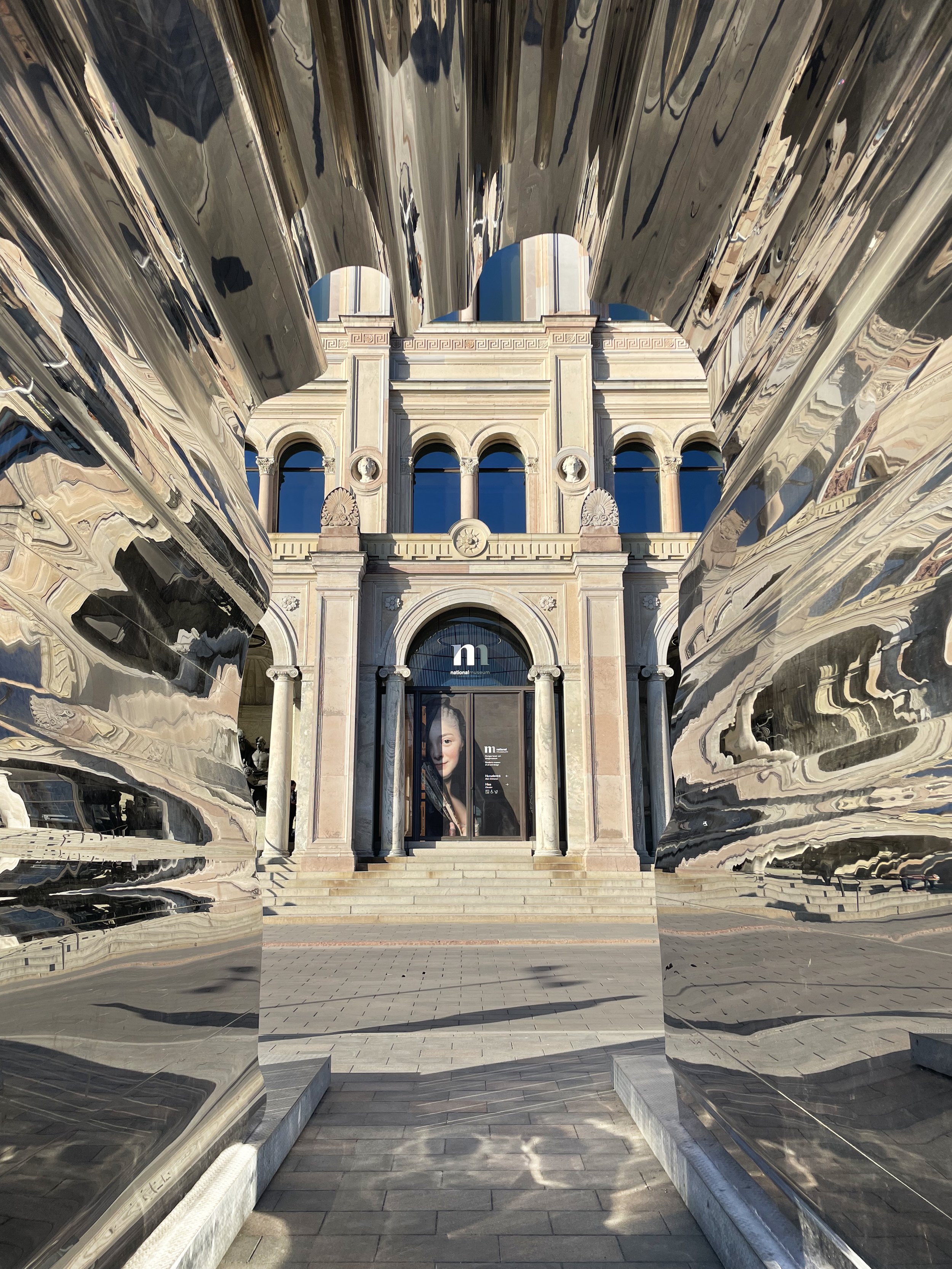 View of a historic stone building with arched windows and stairs, seen through metallic sculptures with a reflective surface. The reflection distorts the building, creating an abstract framing.
