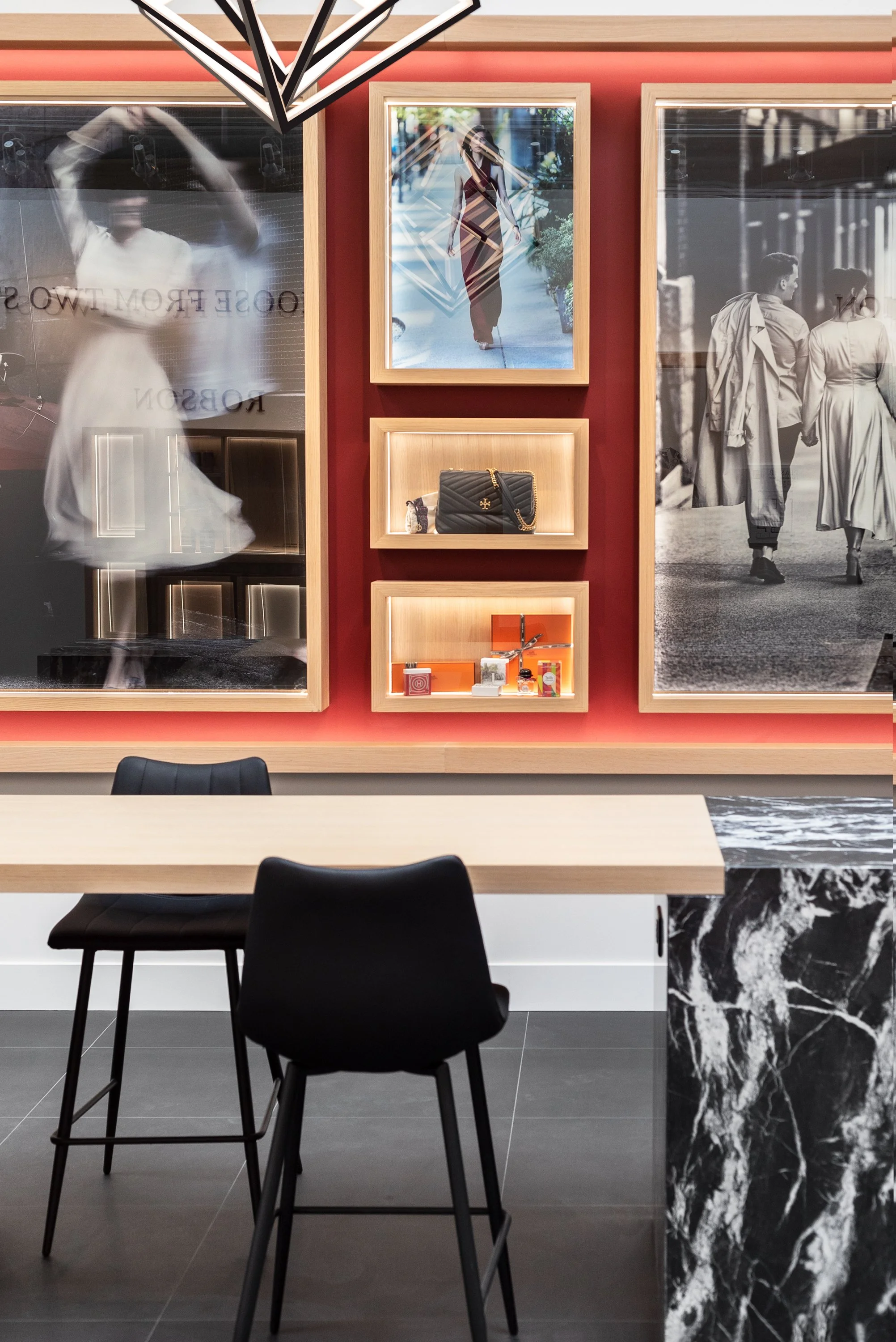 Modern retail store interior with a wooden table and black chairs, decorative wall with framed photographs, handbags, and artwork, and a black marble accent on the table