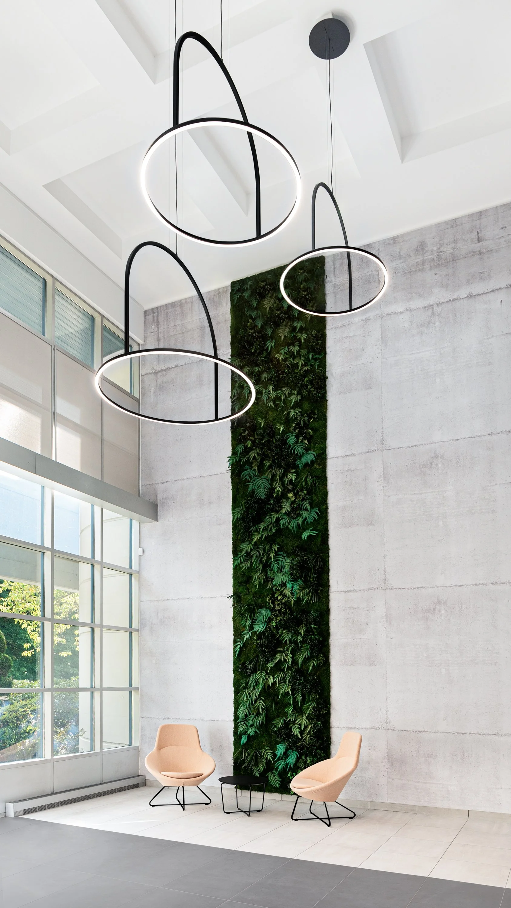 Modern lobby with two beige chairs, a small black table, a green vertical plant wall, and hanging circular light fixtures.