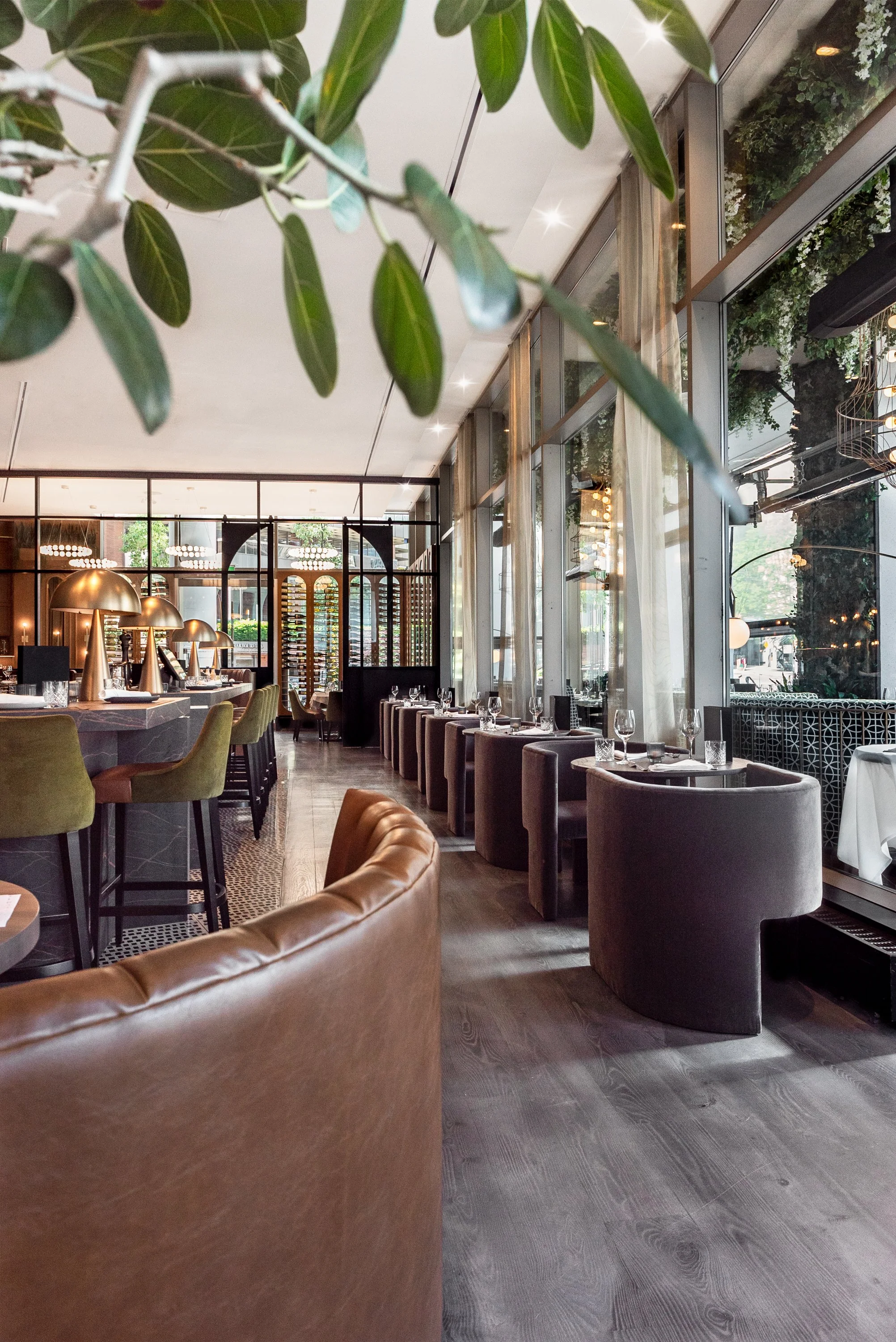 Interior of a modern restaurant with large windows, green and brown chairs, and a row of tables set with wine glasses and napkins.
