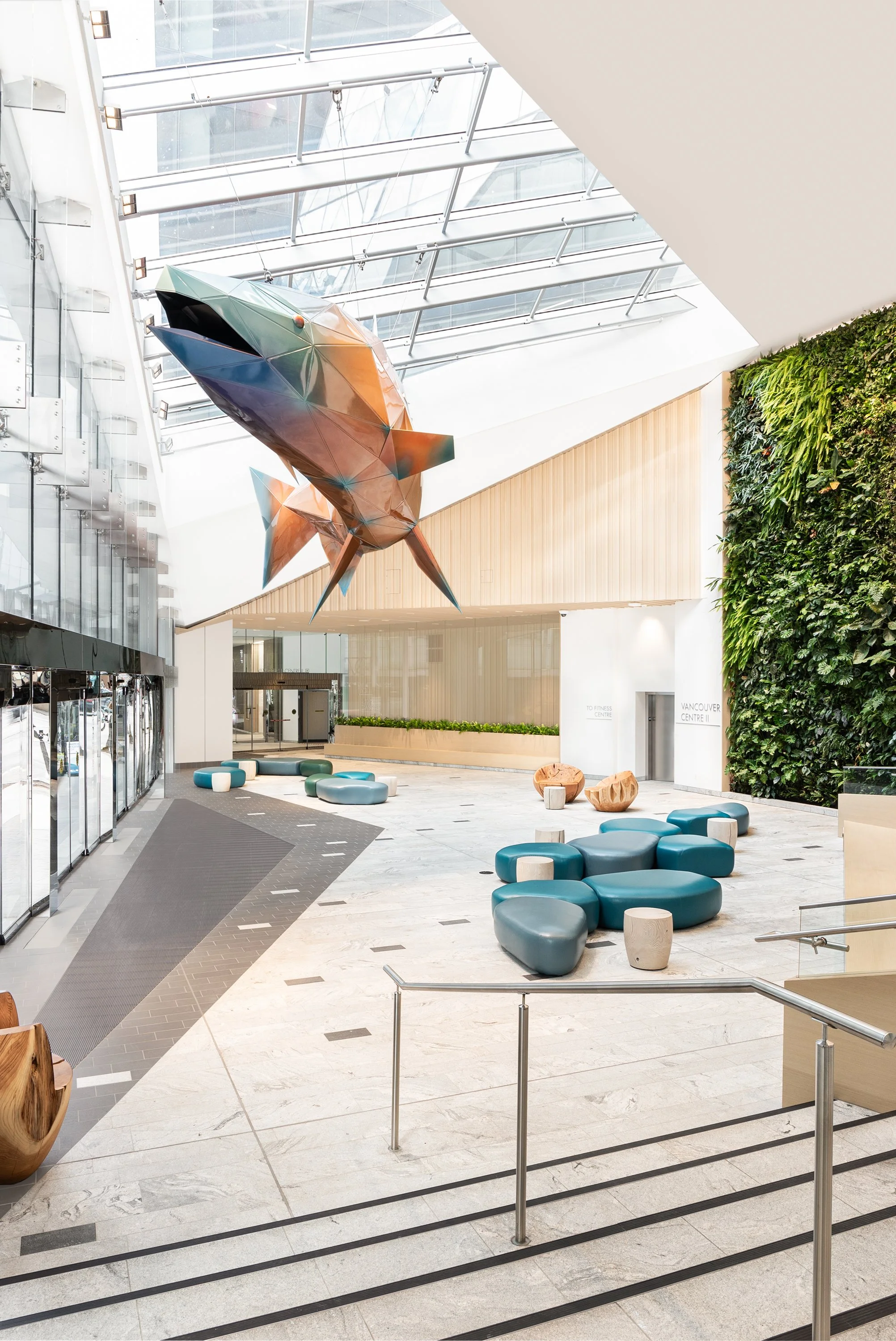 Modern indoor lobby with large glass ceiling, green living wall, colorful abstract fish sculpture hanging from ceiling, and blue and gray seating arrangements.