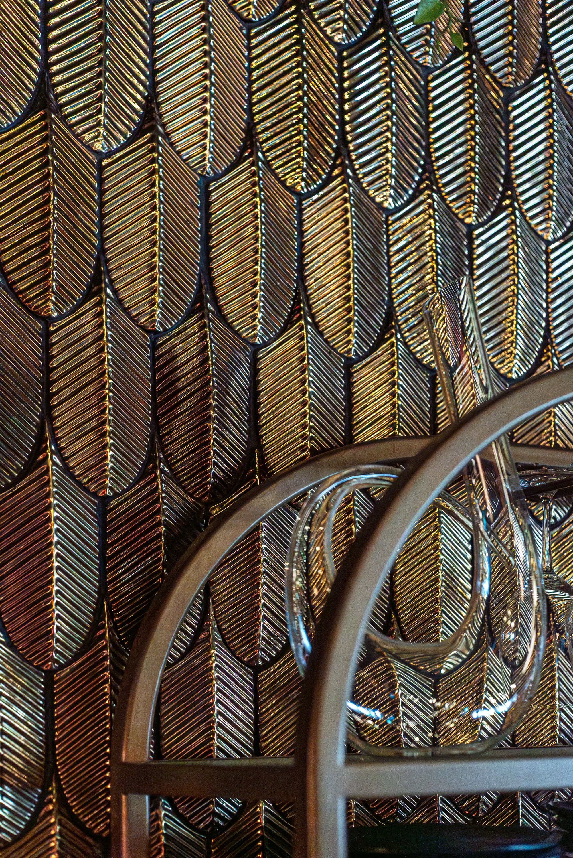 Close-up of a textured wall with a pattern of metallic, leaf-shaped tiles in a gold and bronze hue, reflecting light.
