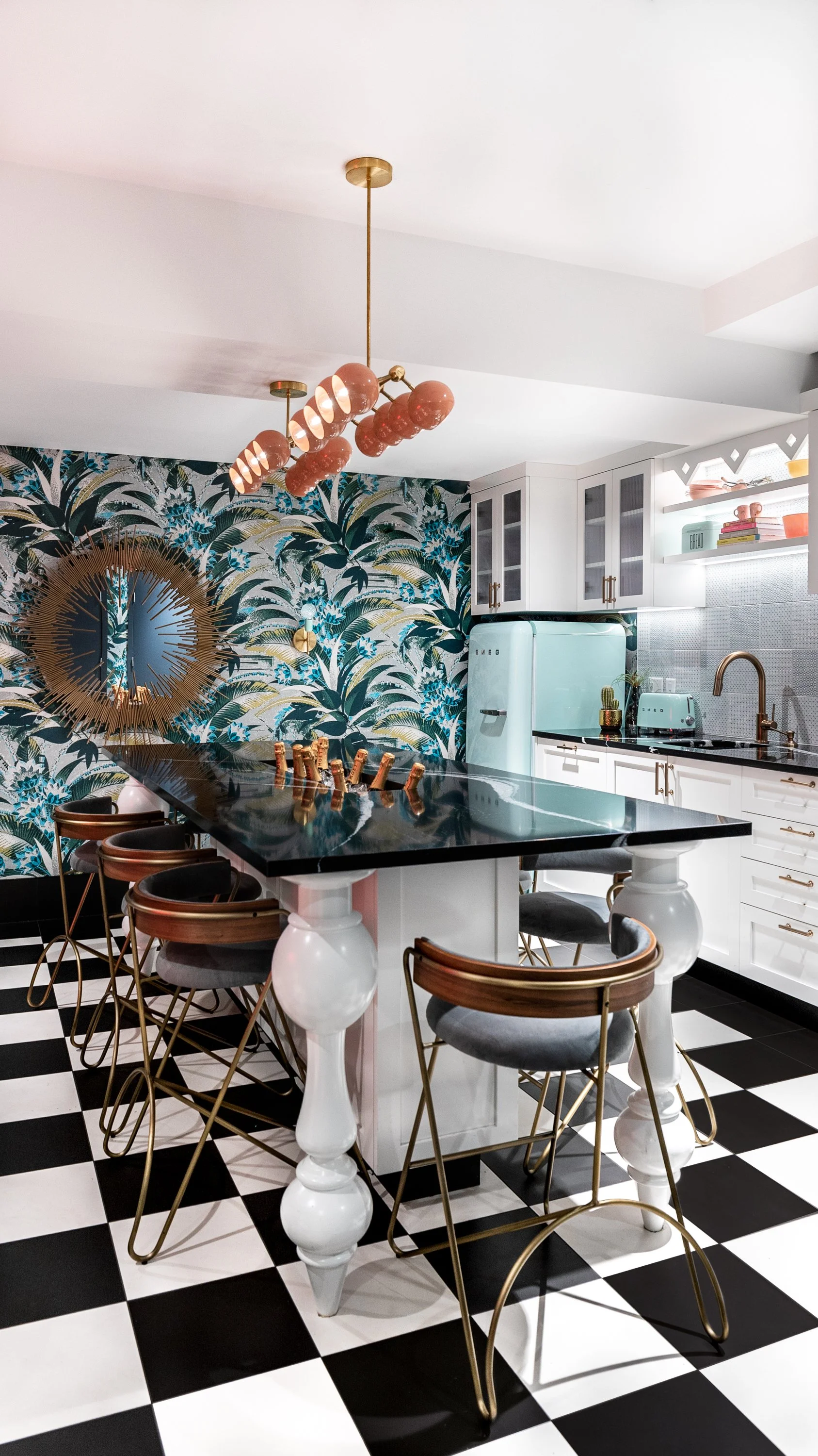 Modern kitchen with black and white checkered floor, white cabinets with gold handles, mint green refrigerator, and tropical wallpaper. Gold bar stools with gray cushions surround a black island with white legs, topped with a glossy black countertop.