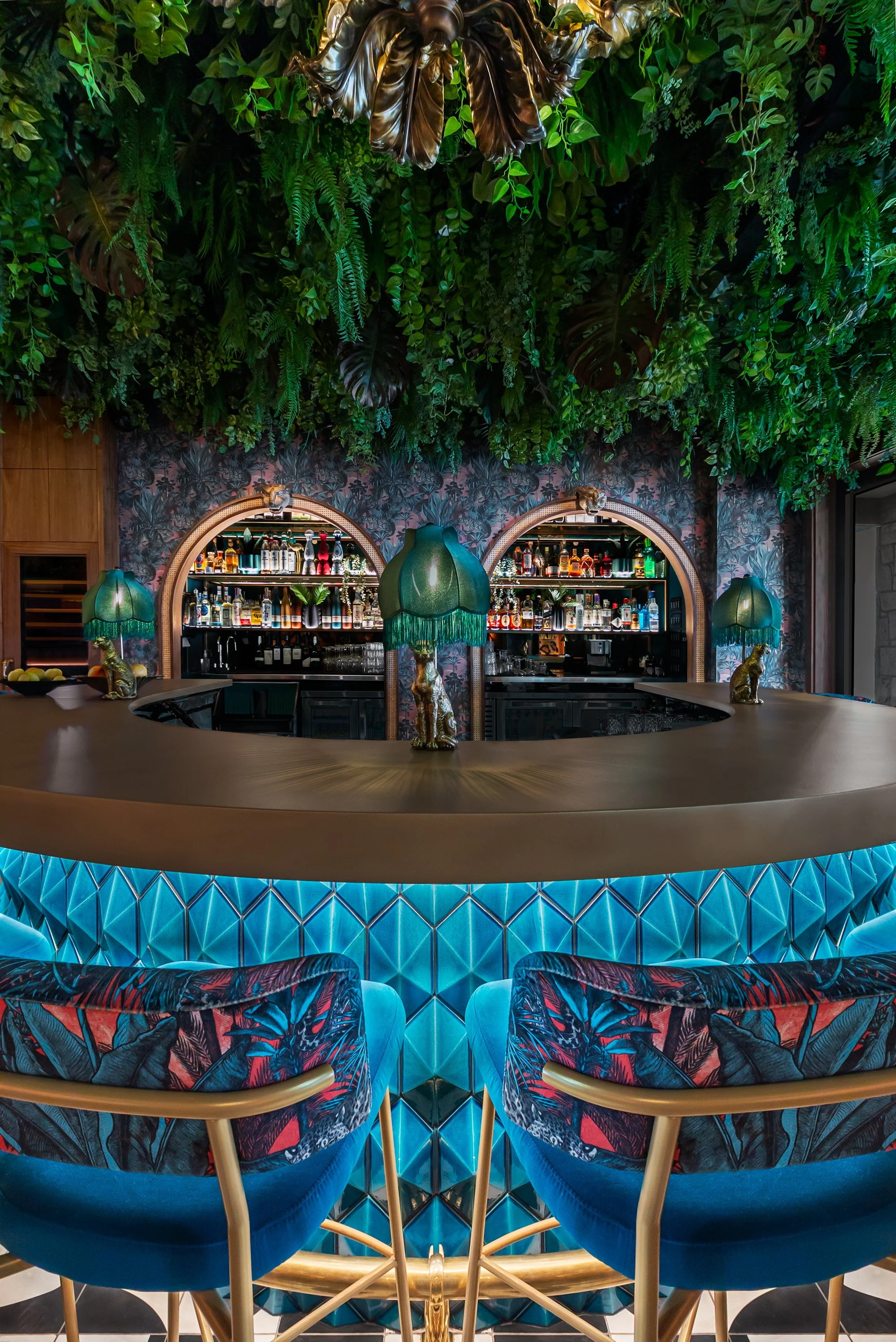 A stylish bar with a curved counter illuminated with blue light, adorned with decorative green lamps and gold animal figurines. Behind the bar, there are shelves filled with bottles, with a lush green plant ceiling overhead and a wall covered in trop