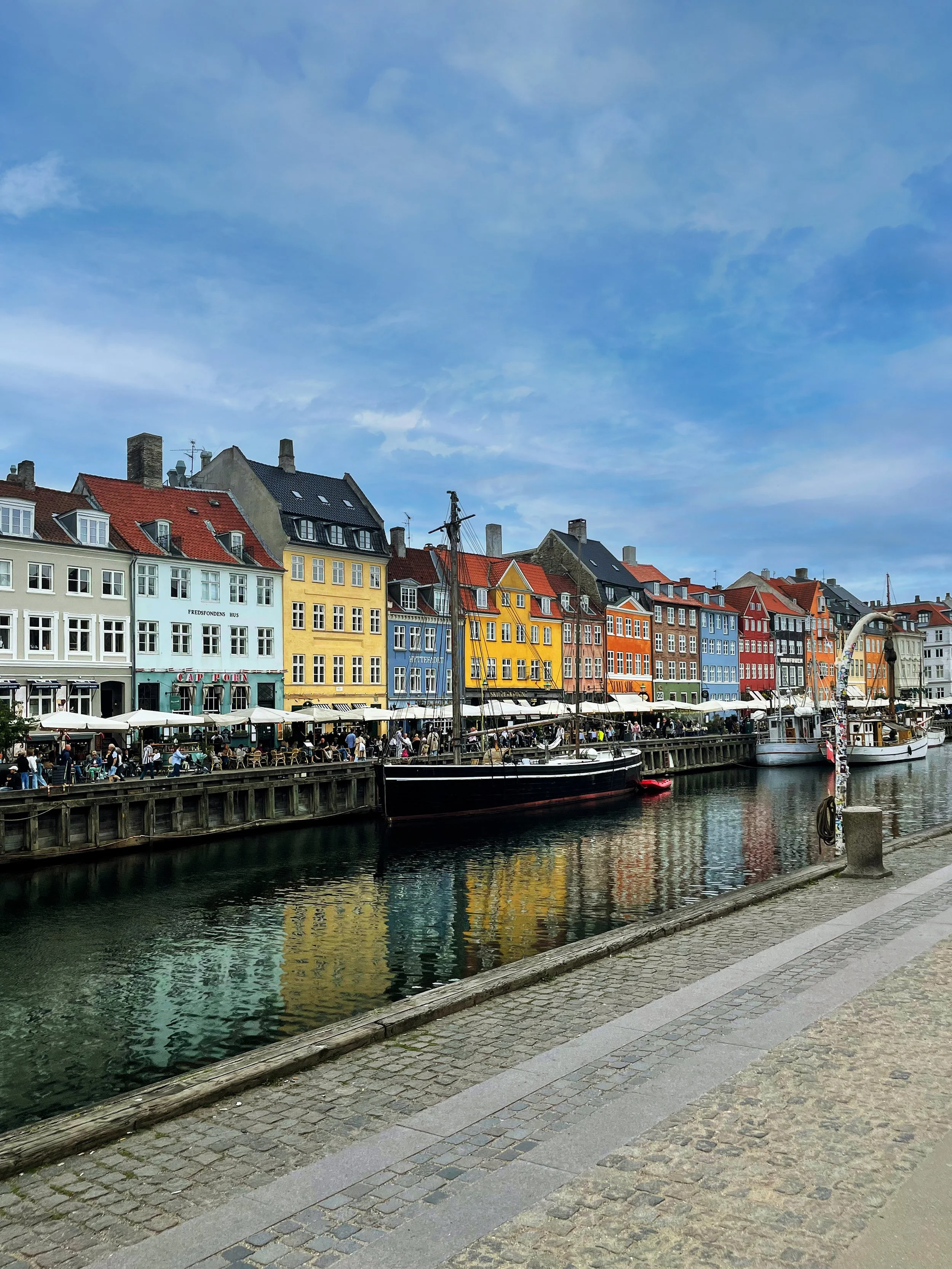 Colorful buildings along a canal with boats docked, and outdoor seating with people, under a partly cloudy sky.