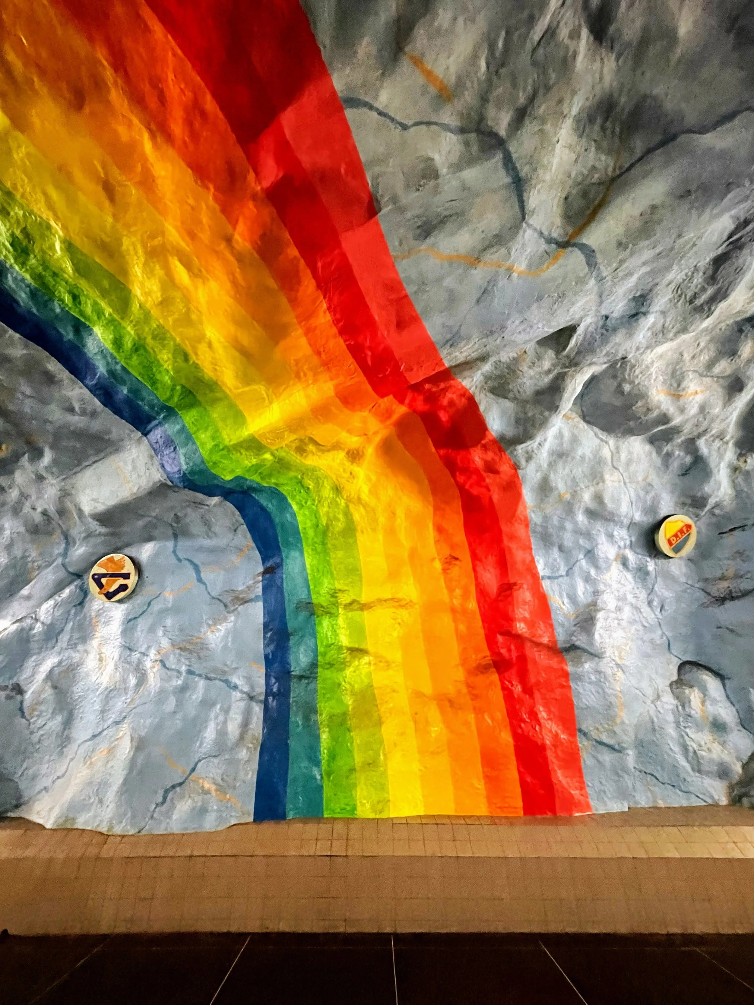 A tunnel with a painted rainbow on the wall, featuring the colors red, orange, yellow, green, blue, and purple, and two symbols on either side at the bottom.