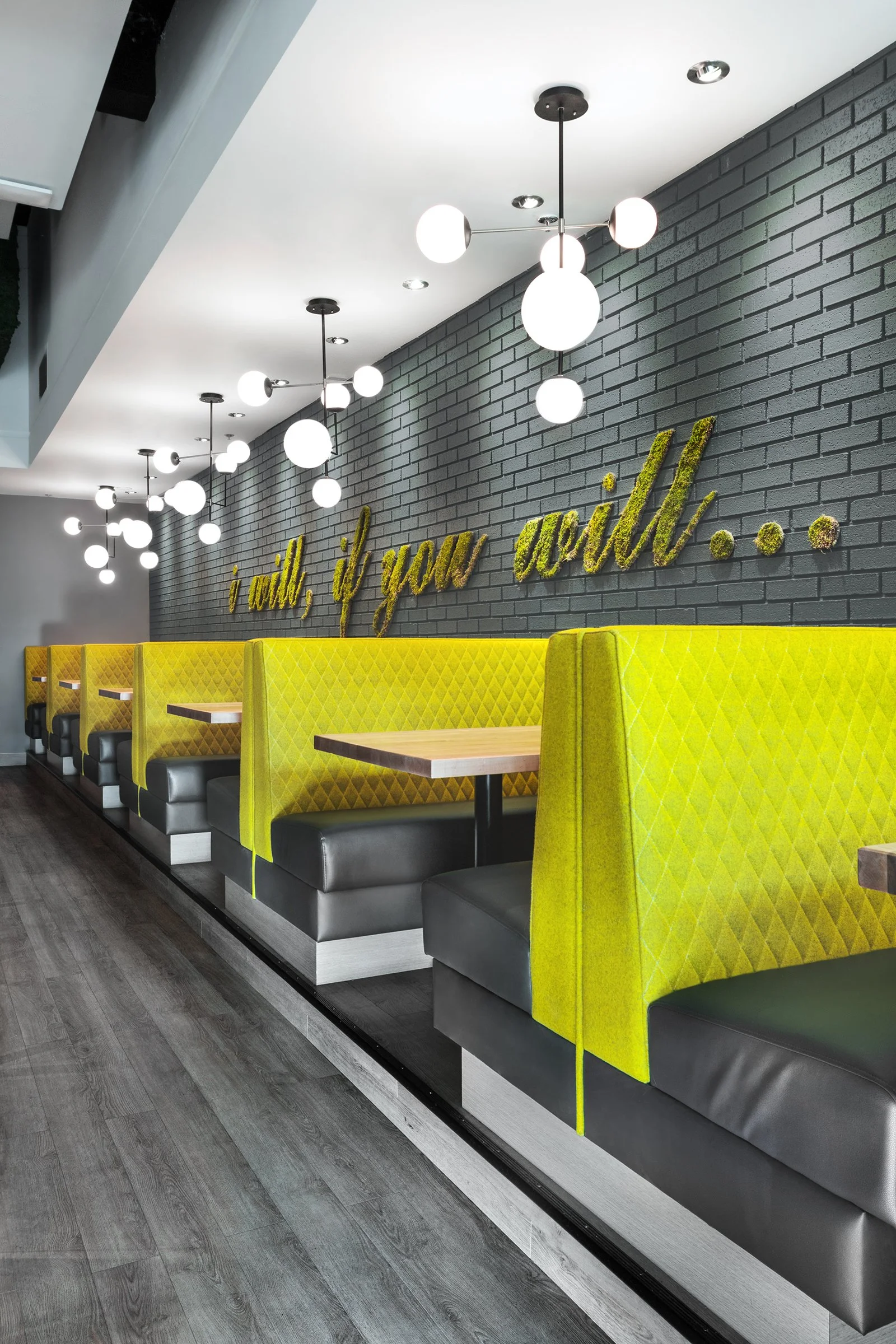 Interior of a modern restaurant booth seating area with yellow padded booths, black cushions, wooden tables, stylish hanging globe light fixtures, dark wooden flooring, and a dark gray brick wall with yellow-green moss-like wall art that reads 'i tol